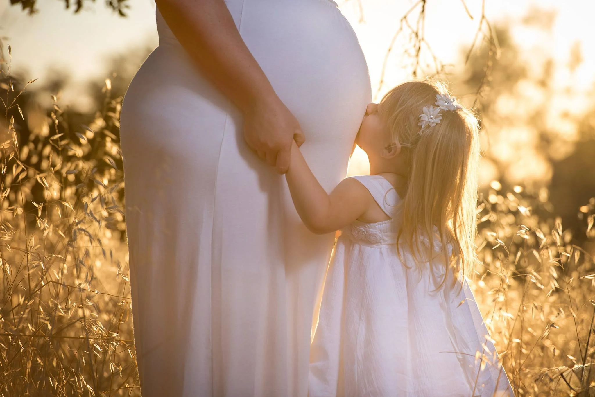 A young girl in a white dress and floral headband is giving a kiss to a pregnant woman in a white dress, standing outdoors with sunlight and tall grass in the background.