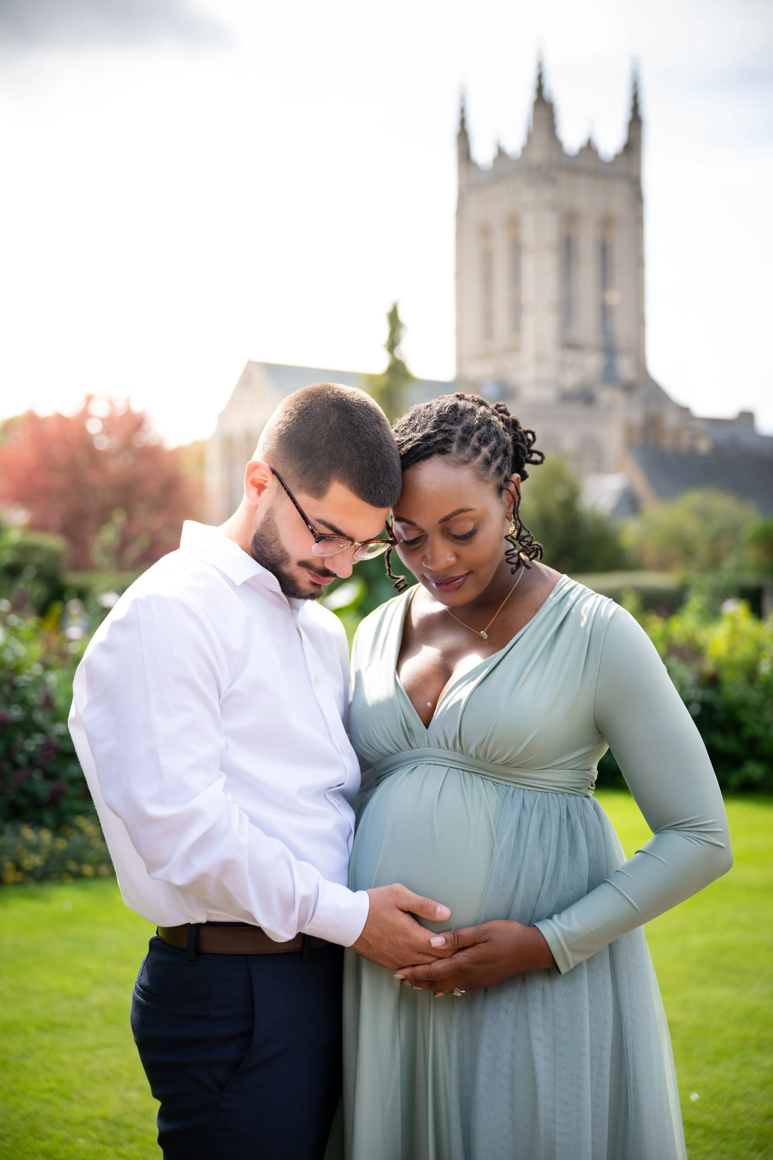 A pregnant woman and a man are standing outdoors on a grassy area, holding her belly and looking down, with a historic church in the background.