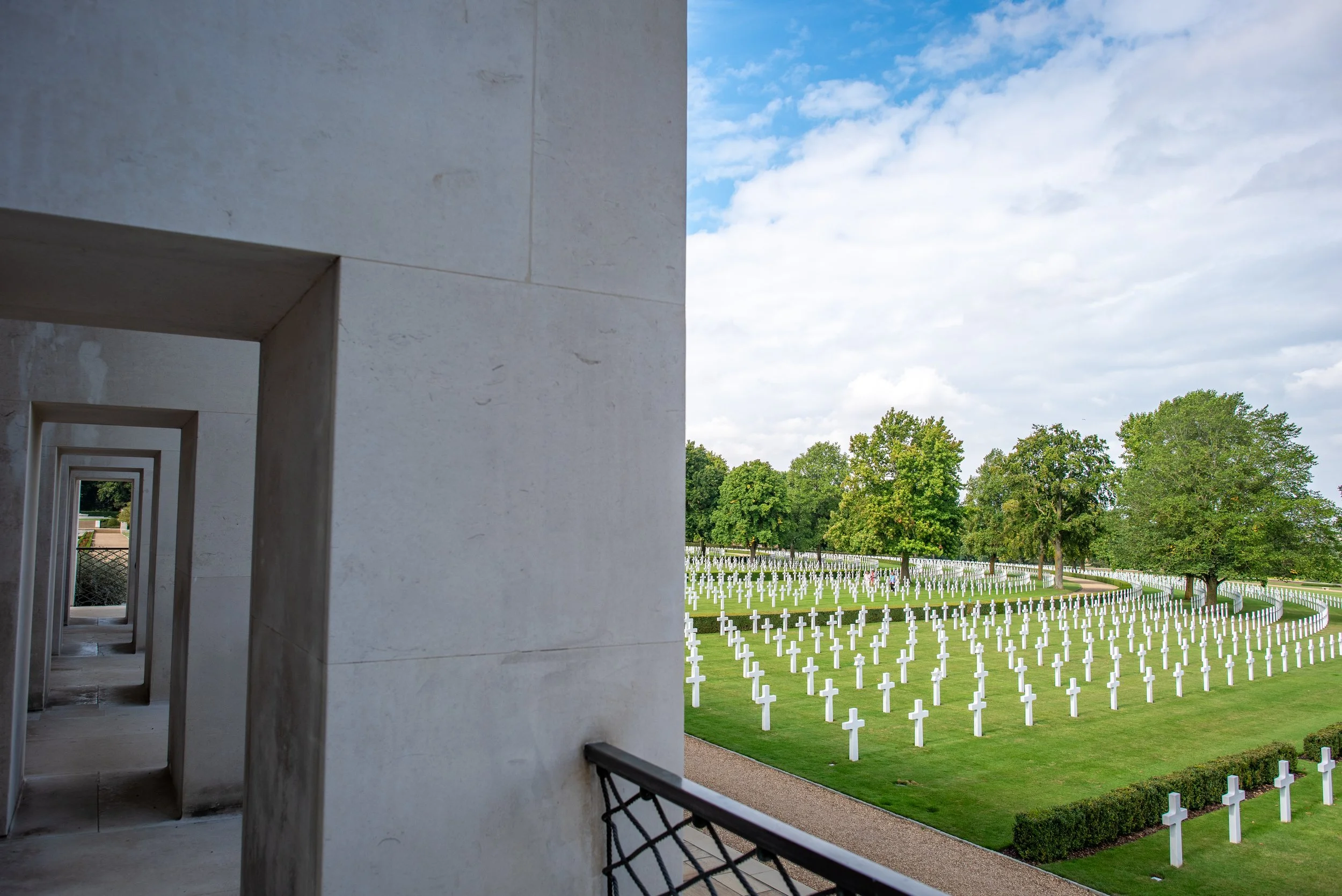View of the American Military Cemetery with rows of white crosses and trees under a partly cloudy sky.