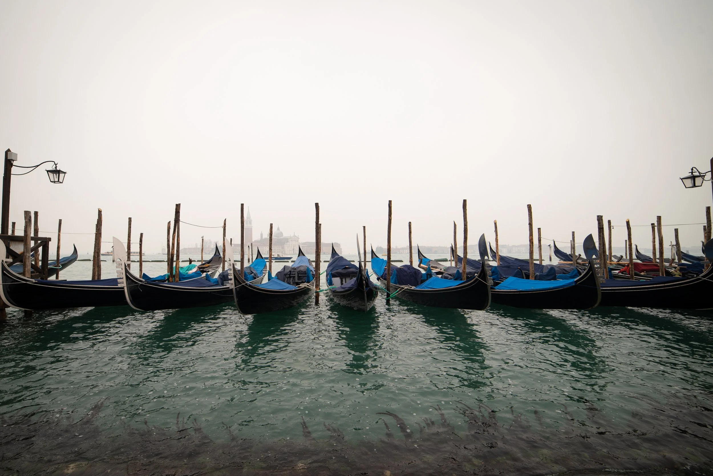 A row of gondolas docked at a Venice waterfront pier, with some buildings visible in the background over the water.
