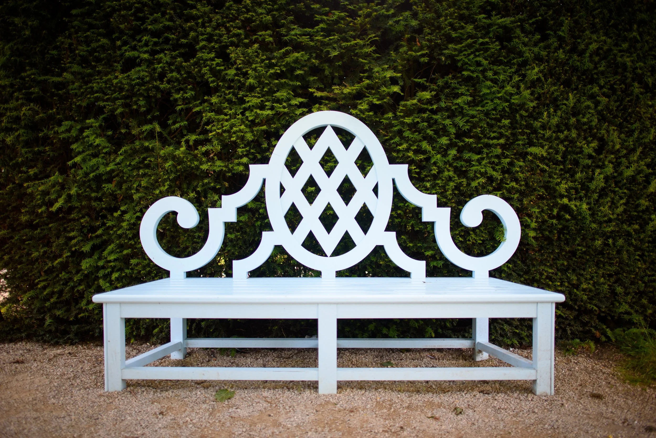 White decorative garden bench with intricate backrest design, set on gravel ground with green hedge background.