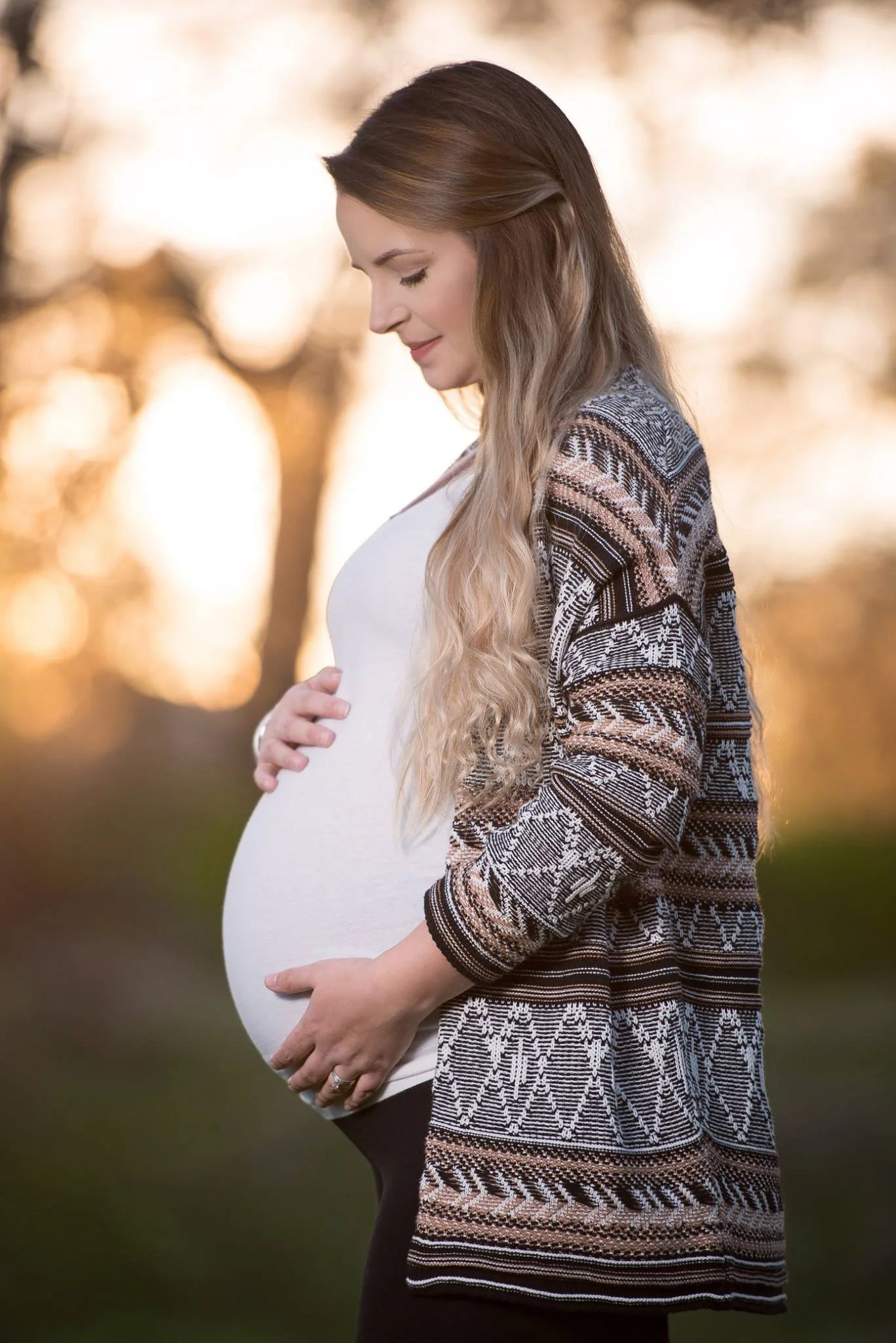 A pregnant woman in profile standing outdoors, cradling her belly, with autumn trees in the background.
