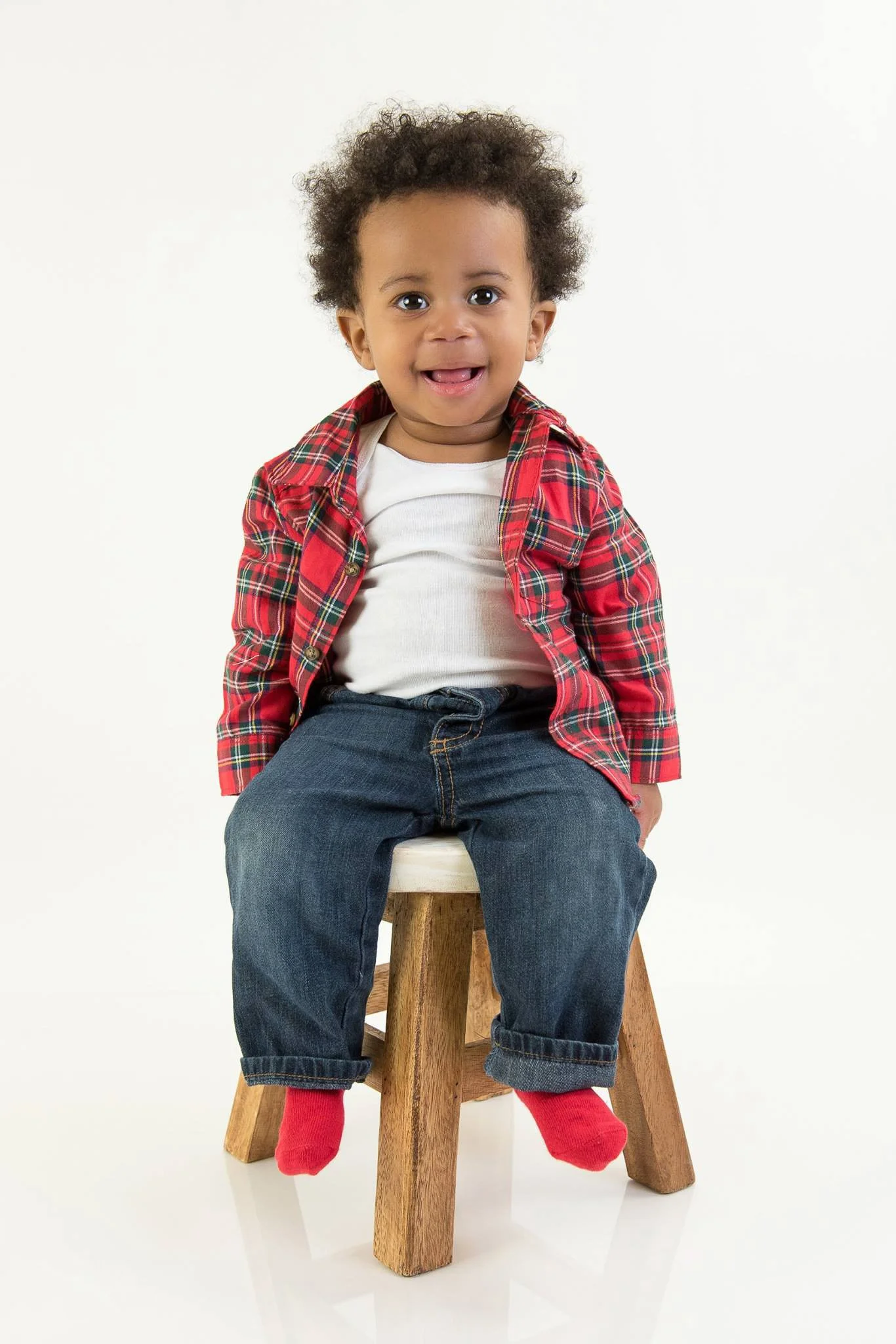 Young boy sitting on a wooden stool, smiling, wearing a red plaid shirt, white t-shirt, blue jeans, and red socks, against a white background.