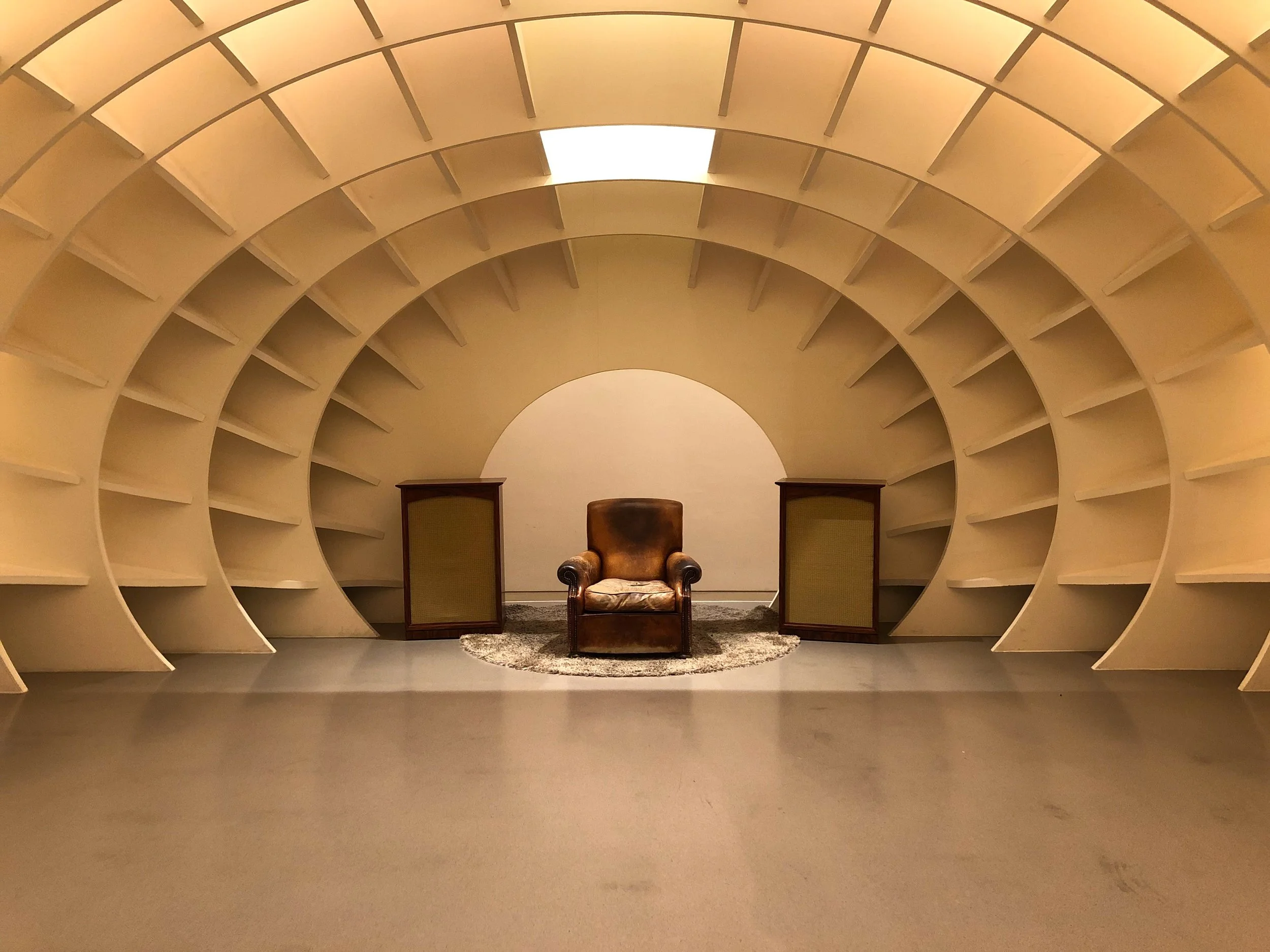 A vintage leather armchair on a small round rug, flanked by two tall cabinets, inside a shell-shaped, cream-colored room with curved, layered walls.