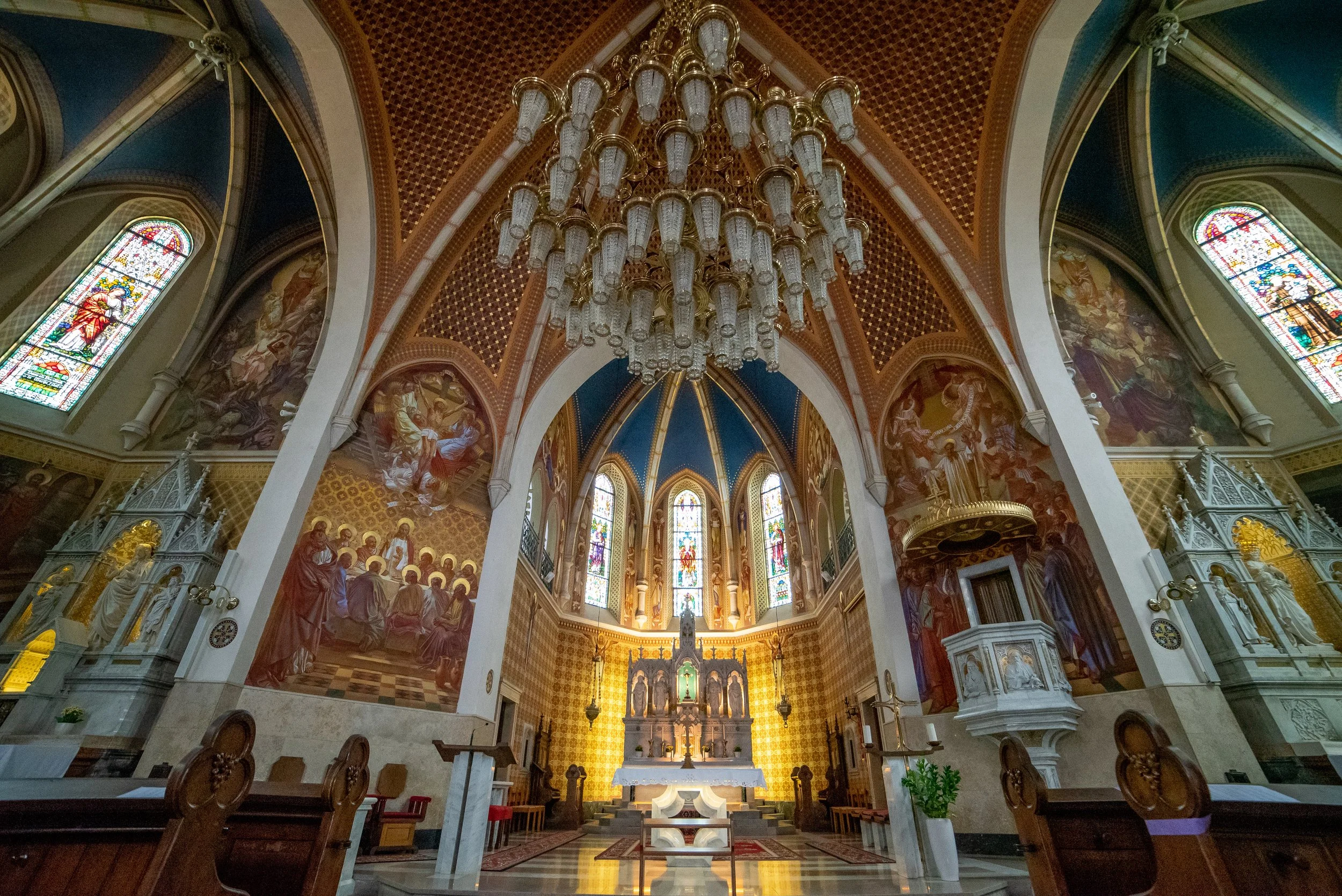 Interior of a church with an ornate altar, stained glass windows, and a grand chandelier hanging from a decorated ceiling.