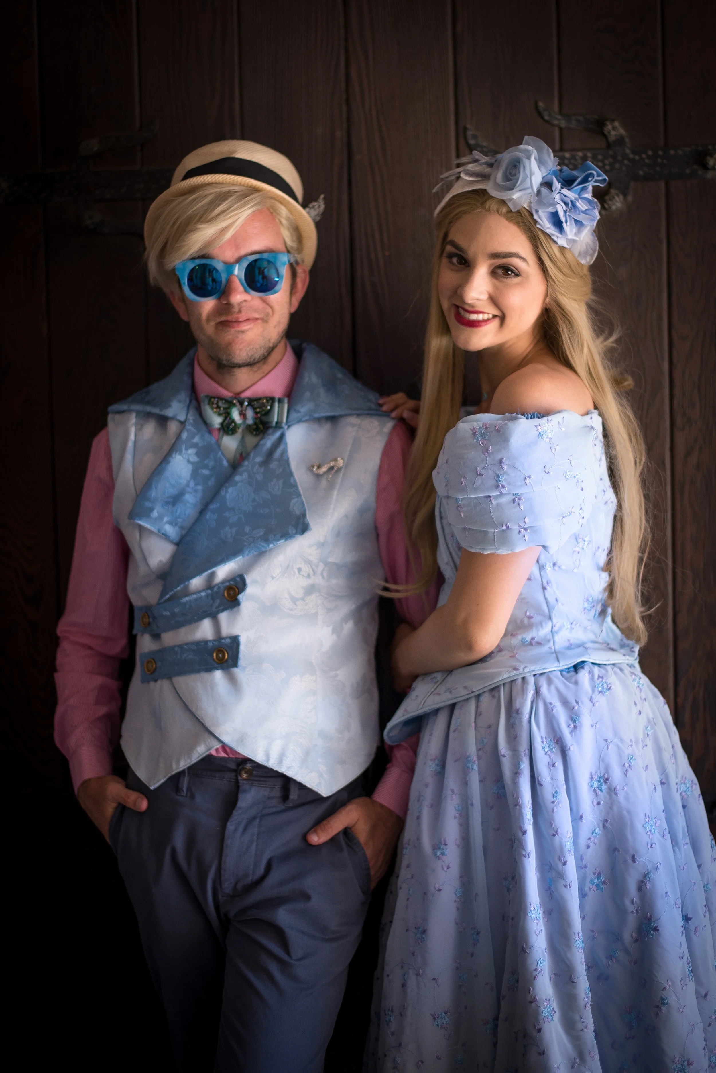Man in vintage-style clothing with blue sunglasses and a hat, standing next to a woman in a light blue floral dress and matching headpiece, both smiling in front of a wooden wall.