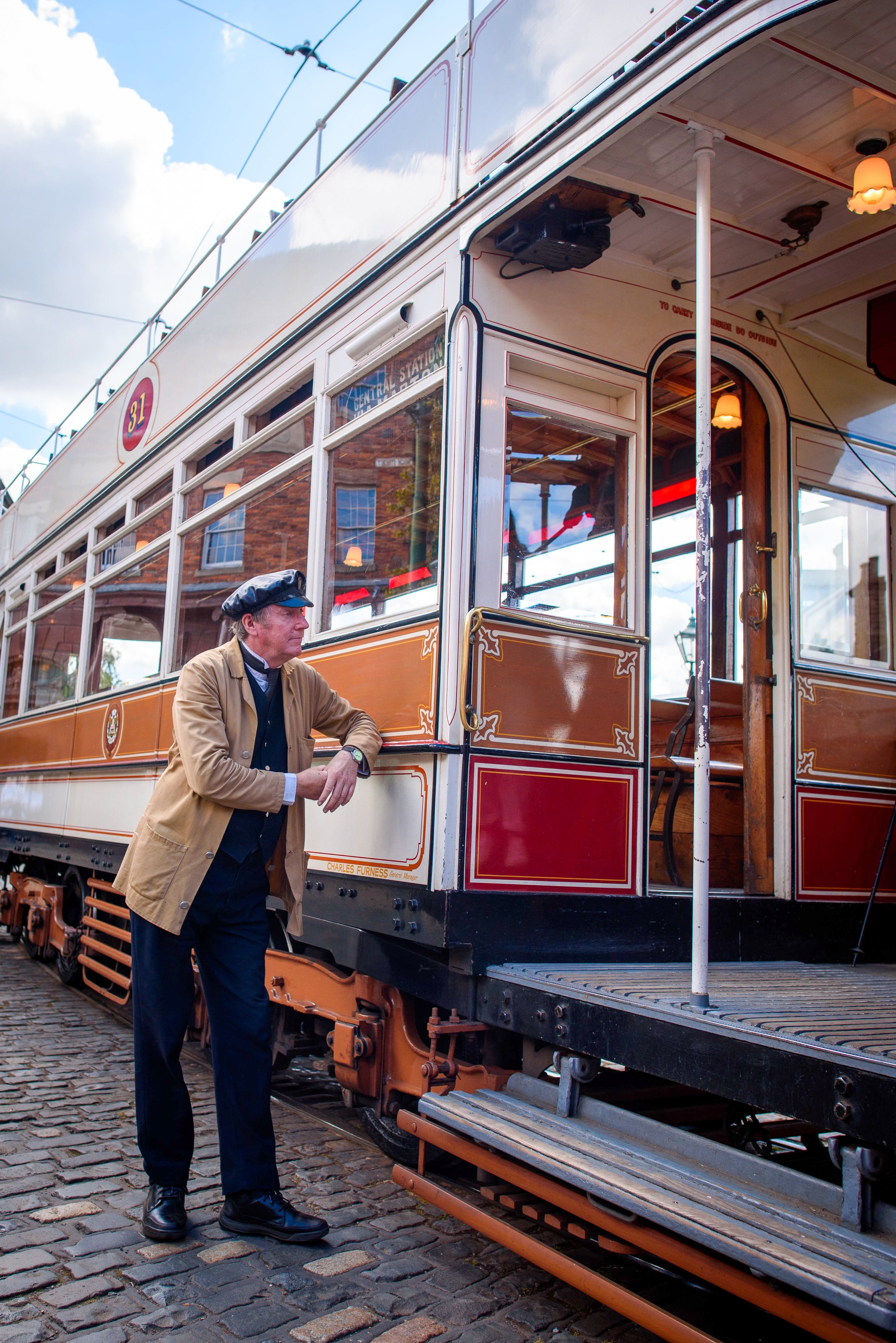 A man dressed in historic clothing leaning on a vintage trolley car, with brick buildings reflected in the windows and a cobblestone street beneath.