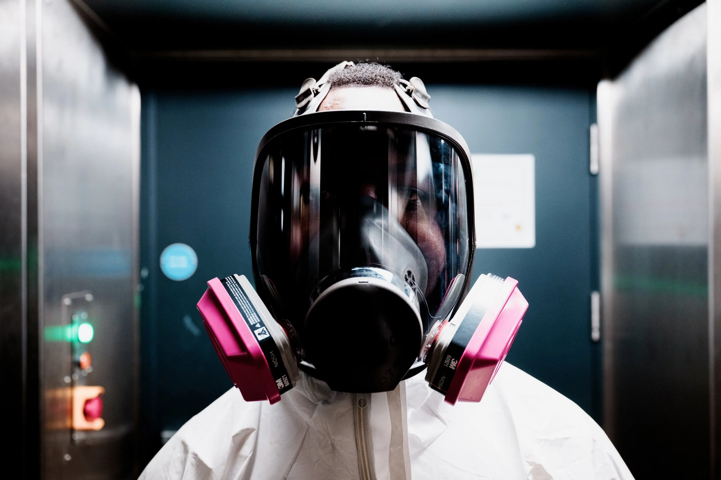 A person wearing a full protective suit, a gas mask with pink filters, and a face shield inside an elevator.