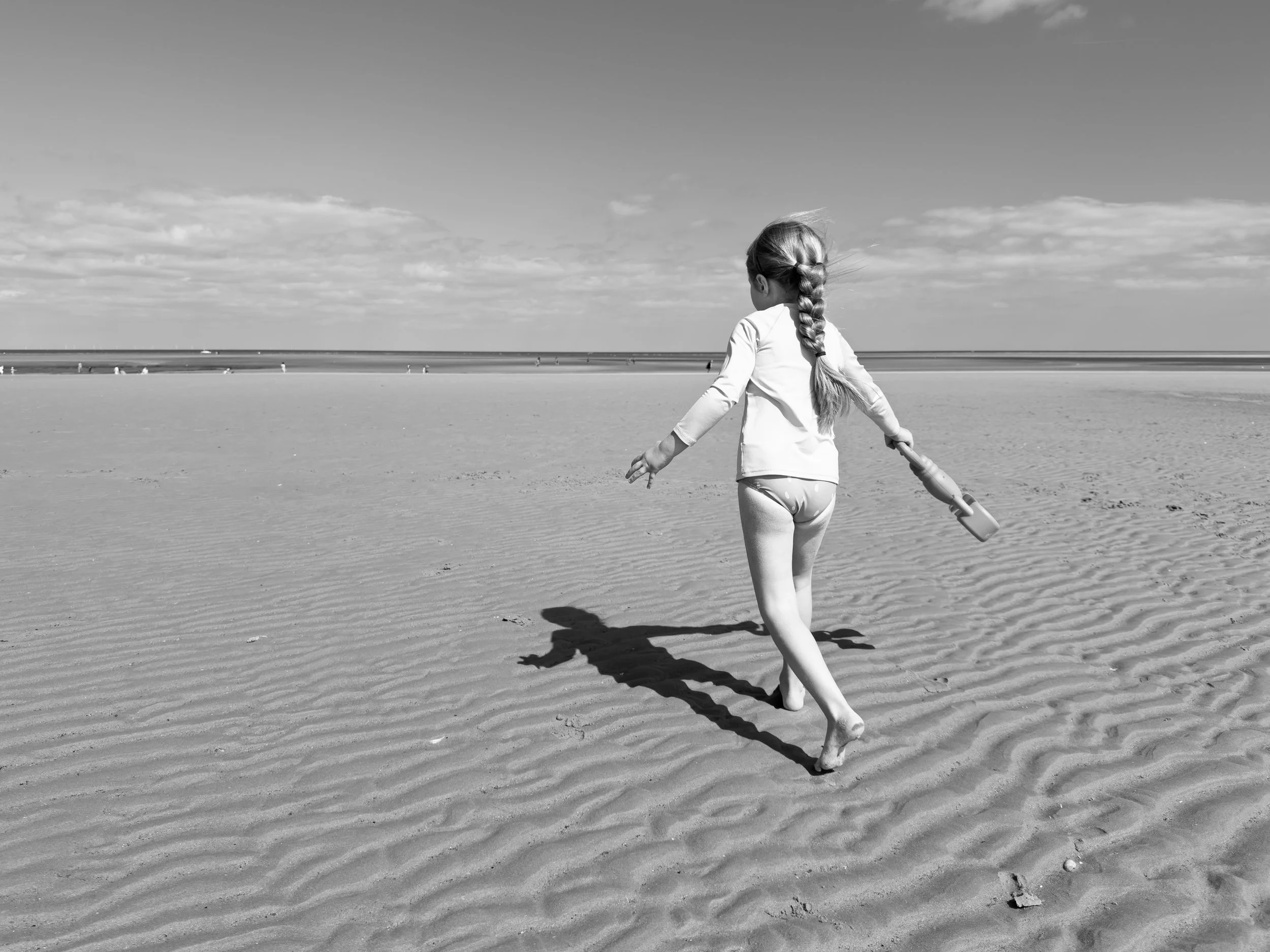 Black and white photo of a young girl walking on a sandy beach holding a small shovel, with her back turned to the camera, long braided hair, wearing a long-sleeve shirt and underwear, casting a shadow on the sand, with the ocean and sky in the backg