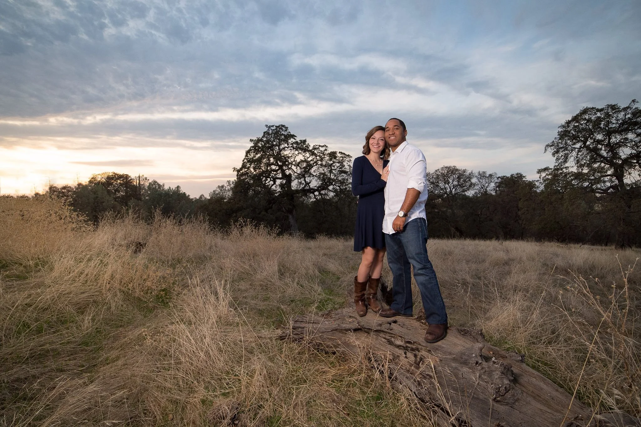 A couple standing close together on a fallen log in a grassy field with trees and a cloudy sky at sunset.