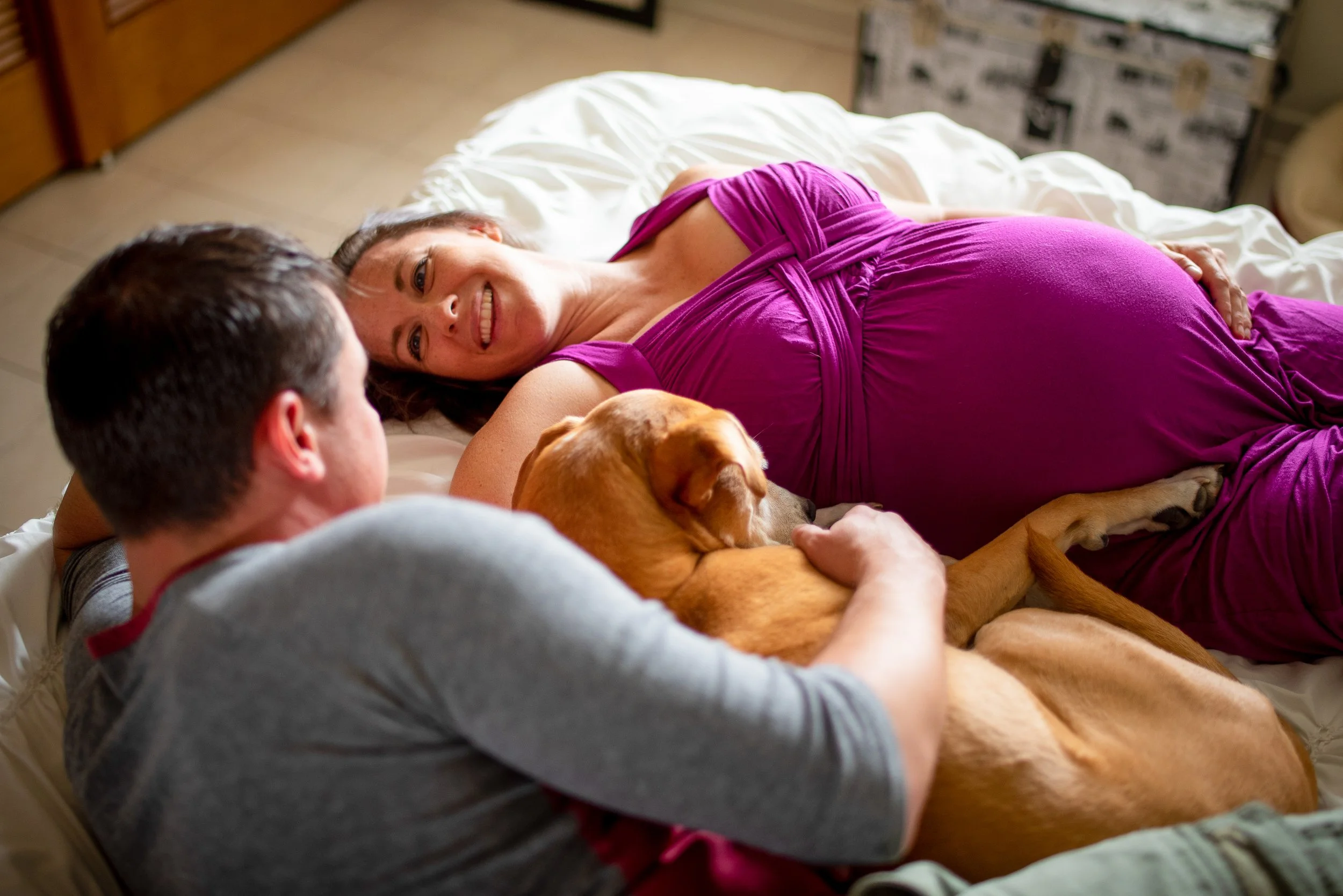 A woman in a purple dress lying on a bed, smiling at a man sitting beside her, with a large brown dog resting on her lap.