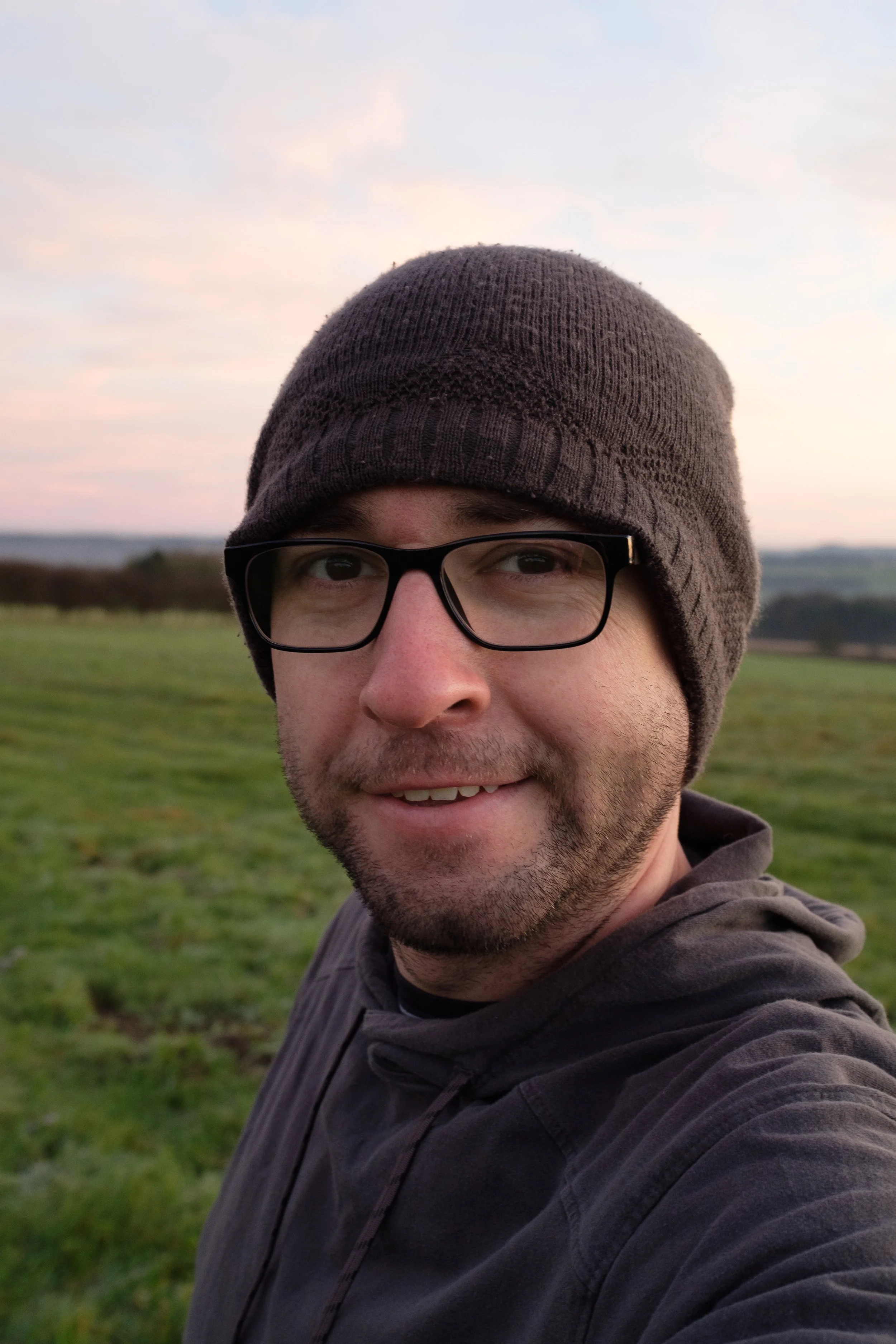 A man wearing glasses and a gray beanie taking a selfie outdoors in a grassy field during sunset.