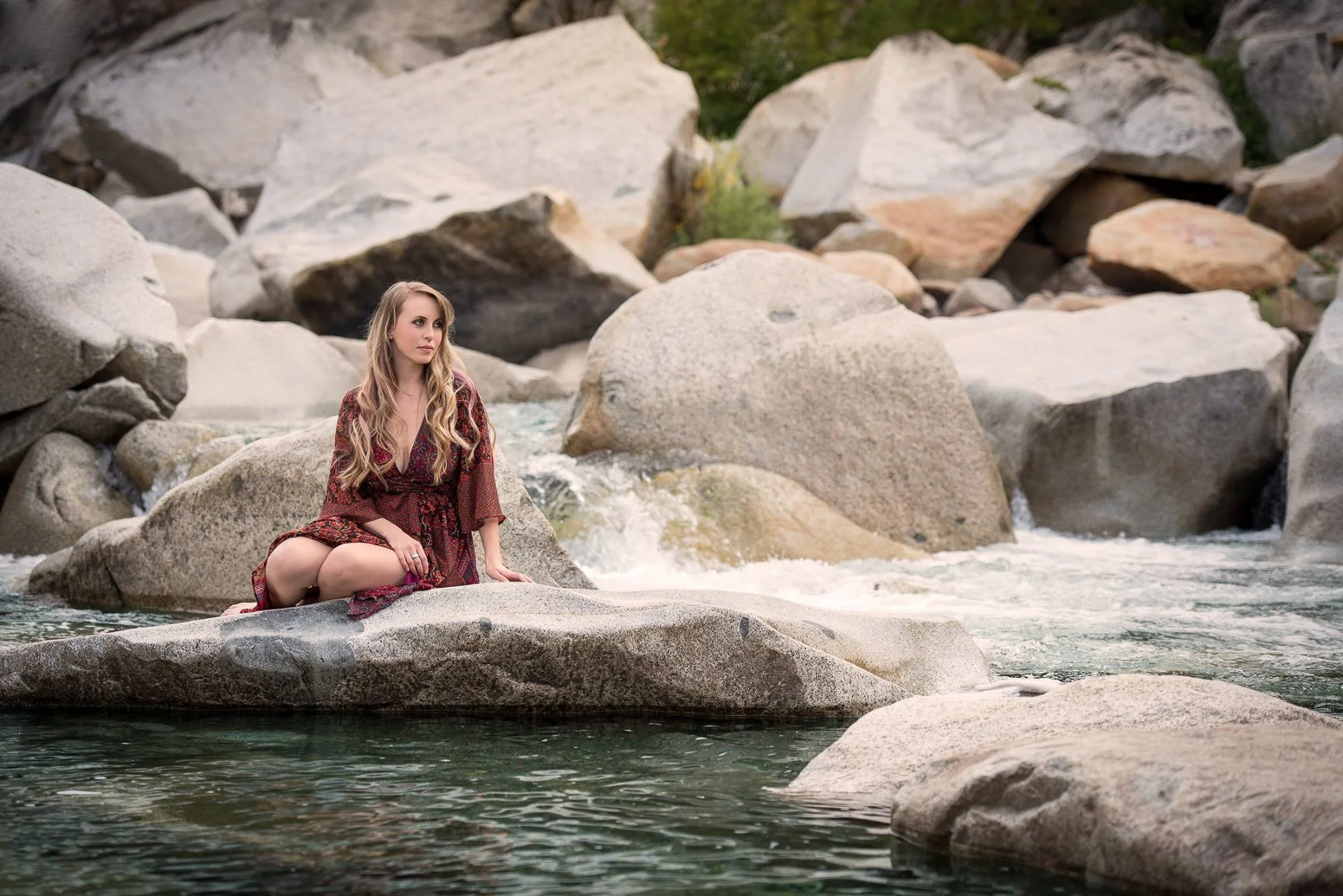 A young woman with long blonde hair, wearing a maroon patterned dress, sitting on a large rock in a river surrounded by many other large rocks.