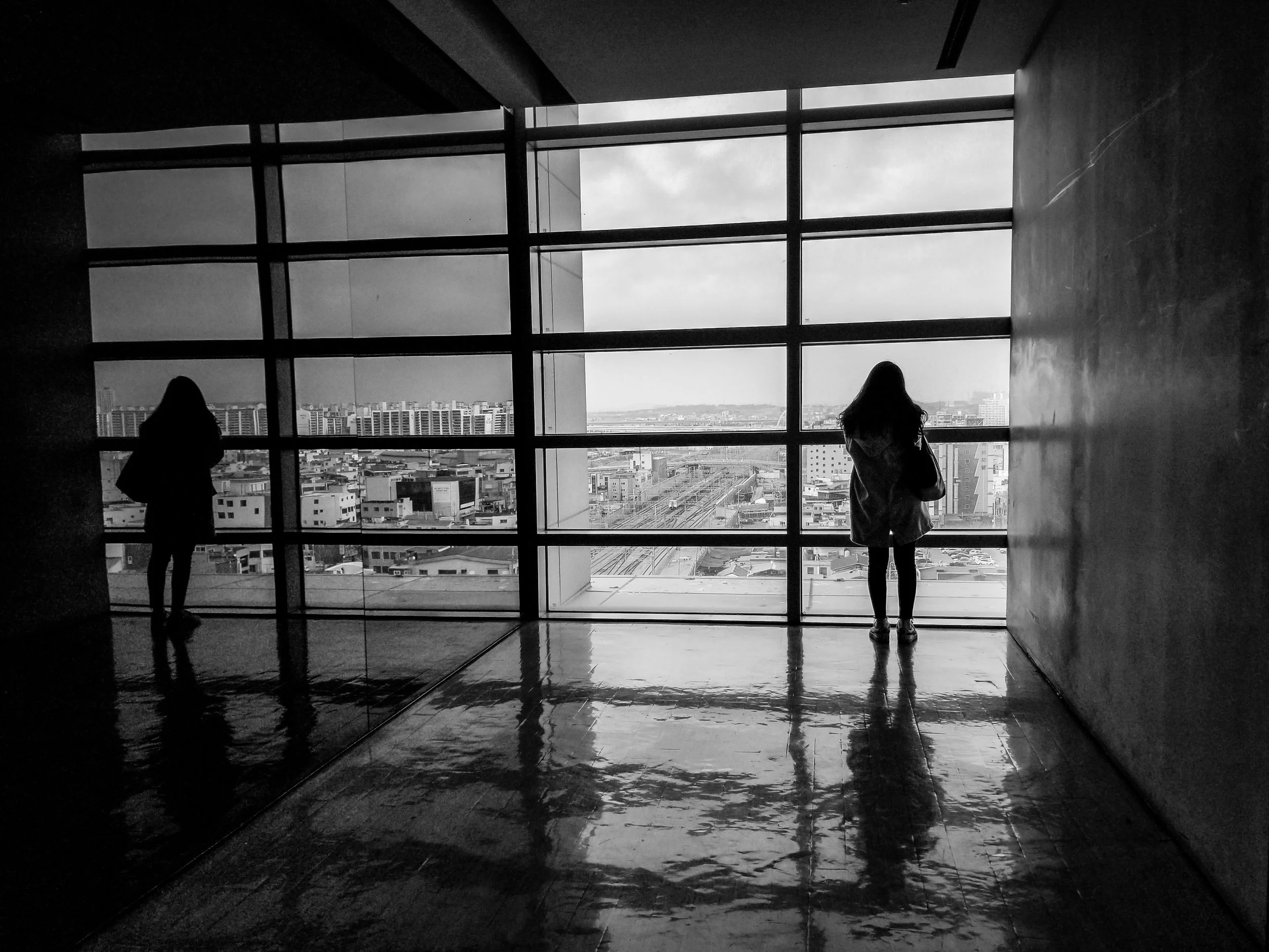 Two women standing near large glass windows overlooking a cityscape, black and white photo.