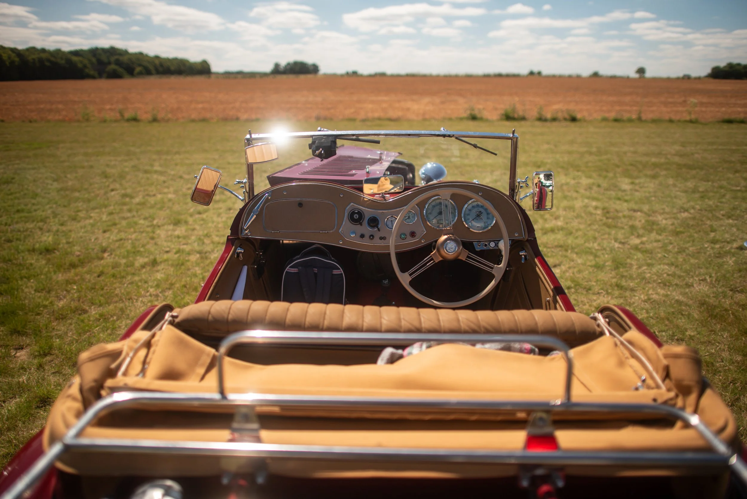 The front interior view of a vintage red convertible car parked on a grassy field with a brown bag in the back seat, and an open landscape with fields and trees in the background under a partly cloudy sky.