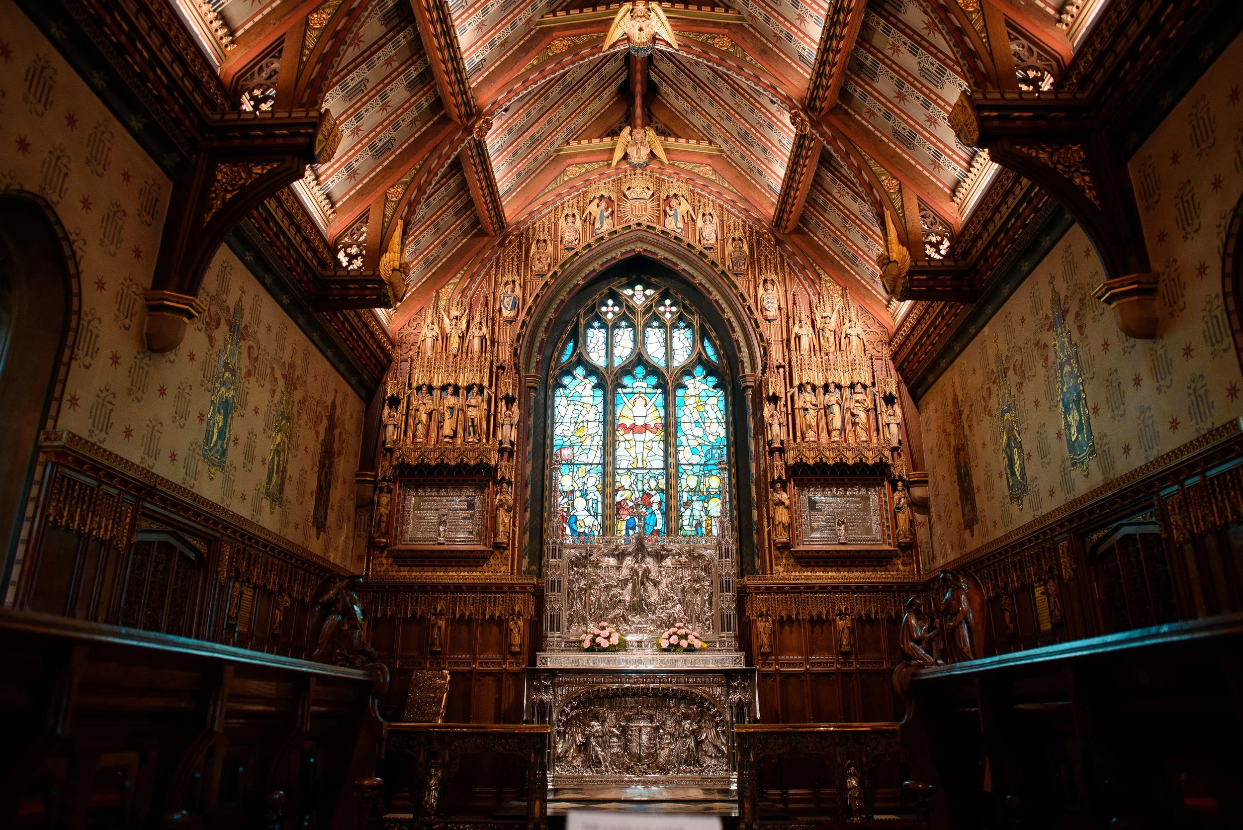 Interior of a church with an ornate altar featuring a large stained-glass window, intricate wood carvings, and religious statues and artwork, with wooden benches in the foreground.