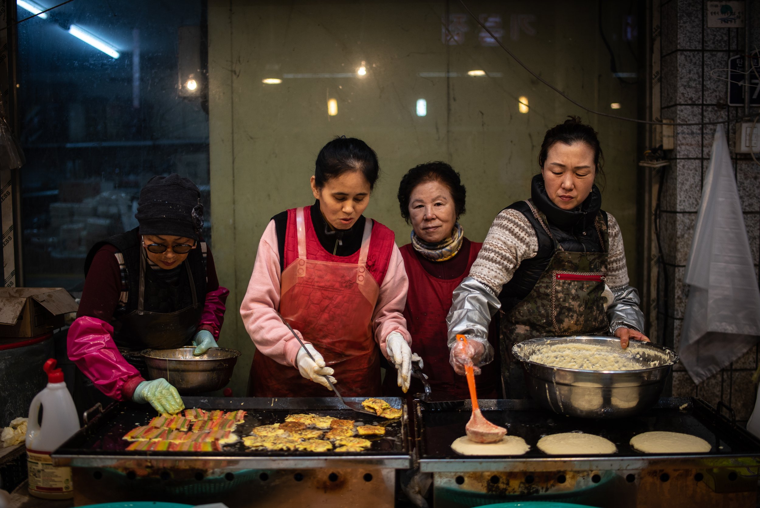 Four women cooking food on a street stall at night, preparing pancakes on griddles.