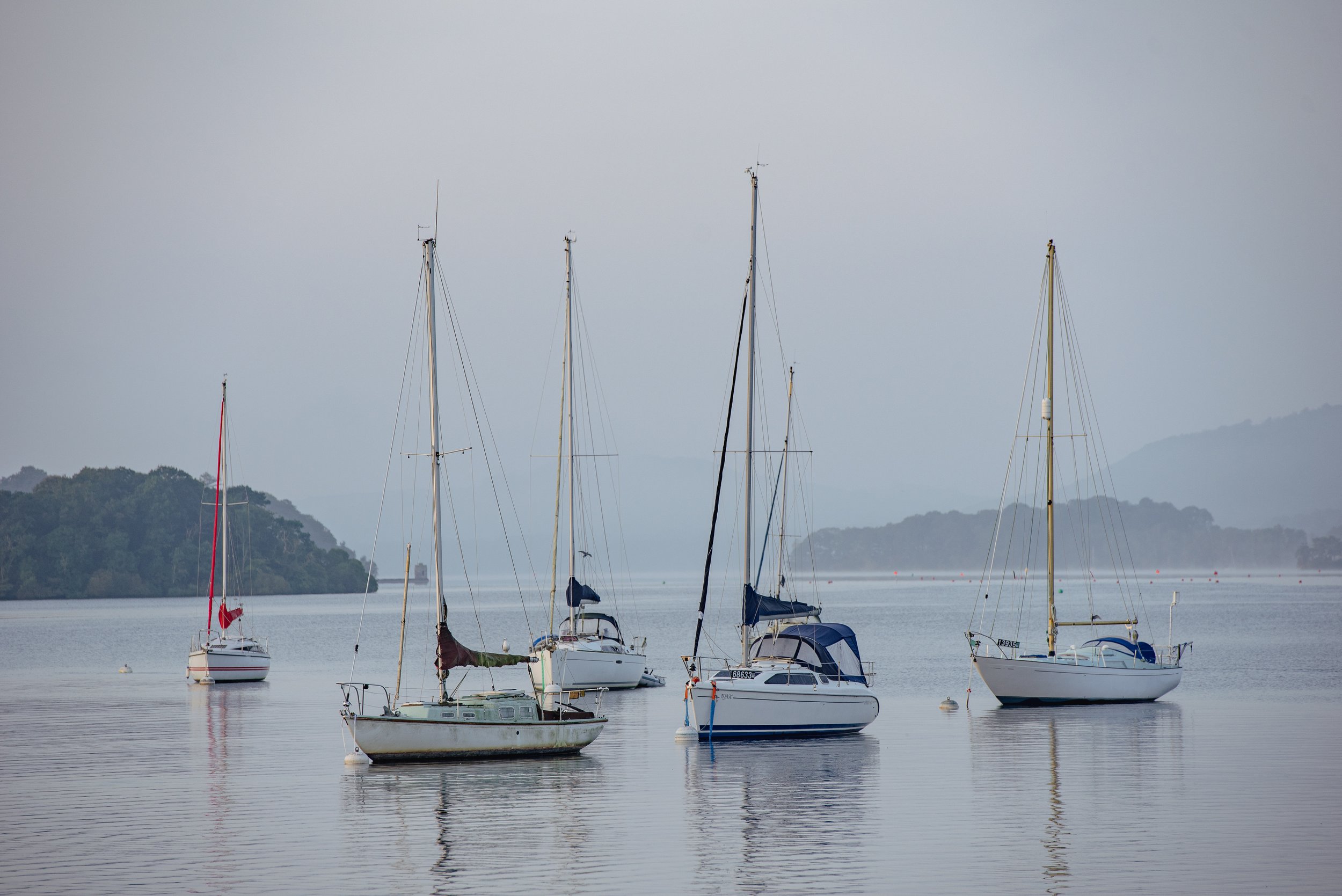 Five sailboats anchored on calm water with forested hills in the background