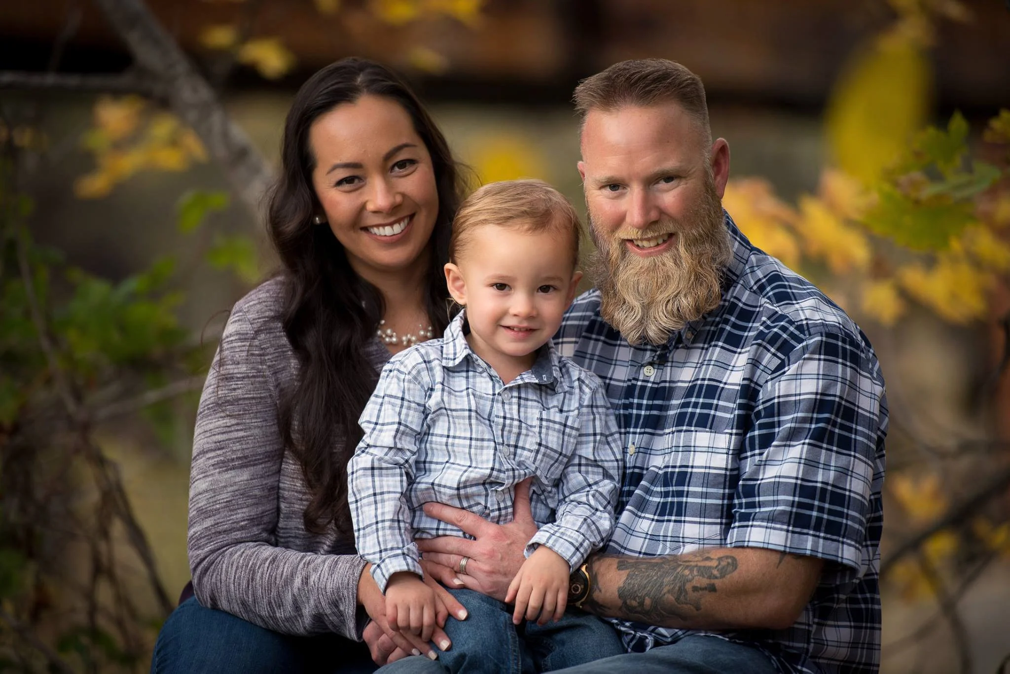 A family of three smiling outdoors during fall, with trees and yellow leaves in the background. The woman has long dark hair, the man has a beard, and the young boy has short light hair.