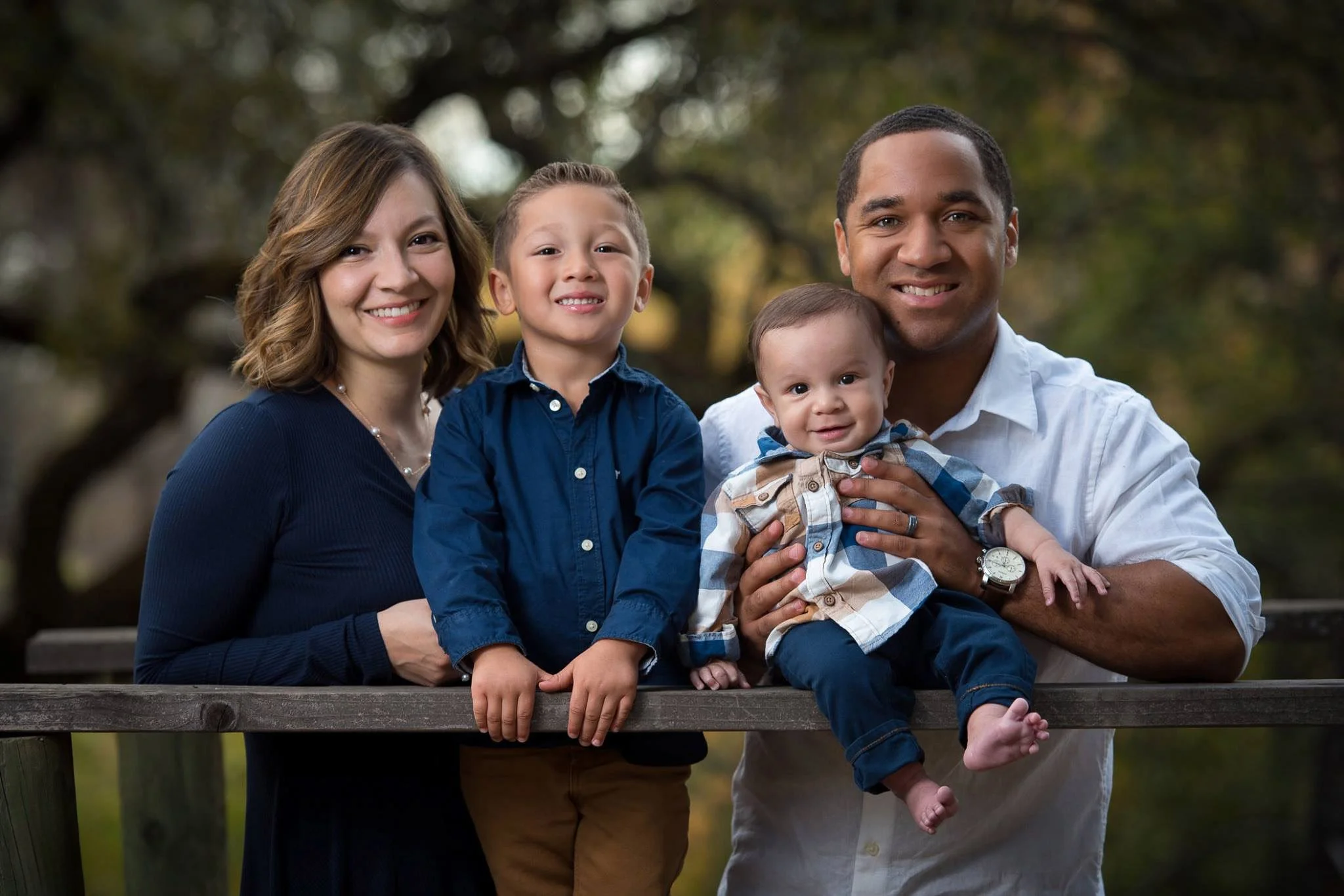 A family of four outdoors, standing behind a wooden railing. The mother has shoulder-length brown hair and is wearing a dark blue dress. The father has short black hair and is wearing a white shirt, holding a baby. The older boy has short light brown