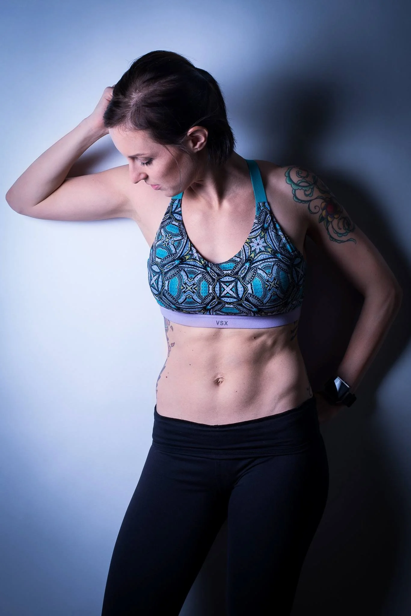 A fit woman with short dark hair, wearing a colorful sports bra and black leggings, standing against a plain wall, with her left hand on her head and her right hand behind her back.