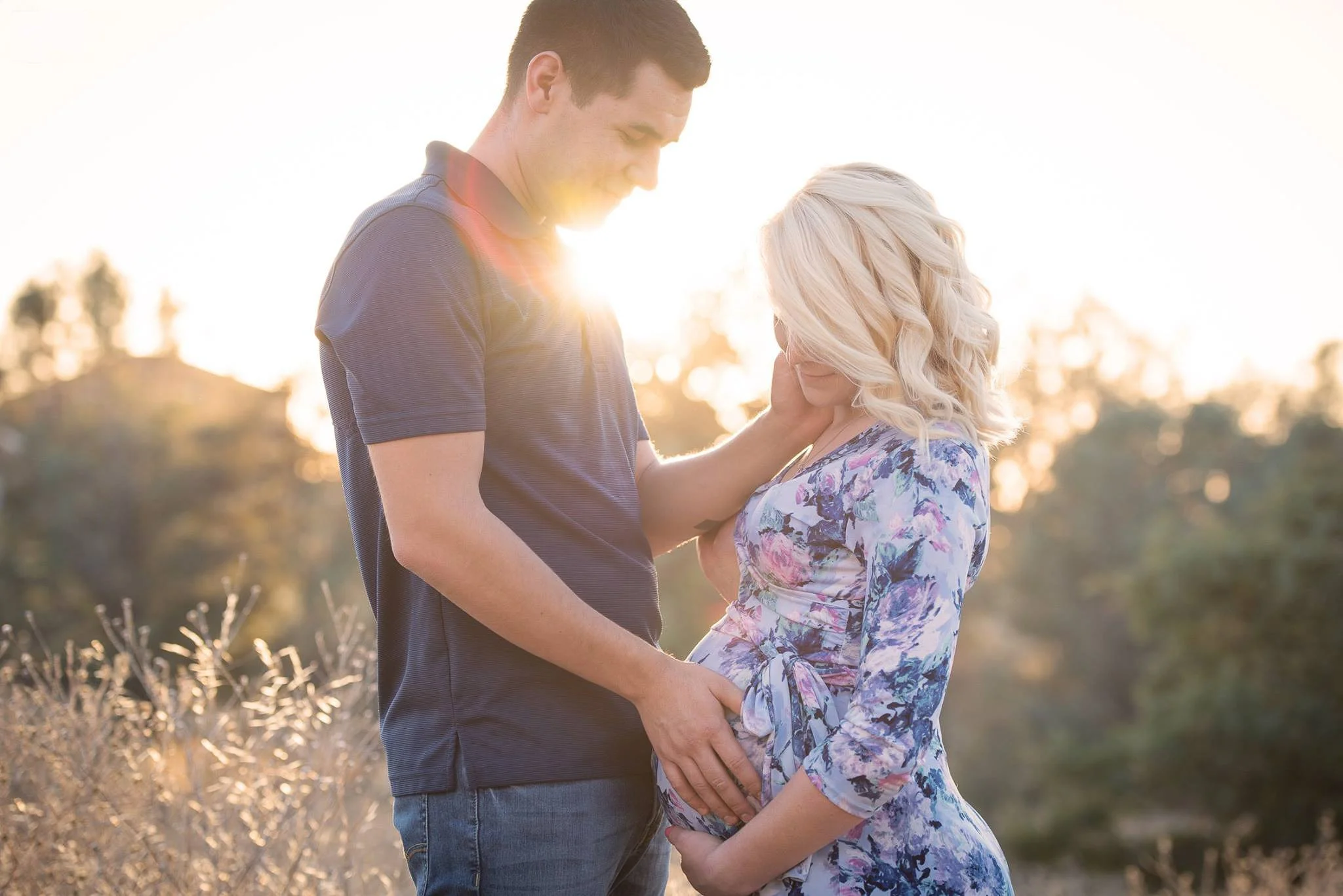 A couple standing outdoors at sunset, with the man gently touching the woman’s pregnant belly and the woman cradling her belly. The man is wearing a navy polo shirt, and the woman is in a floral dress.