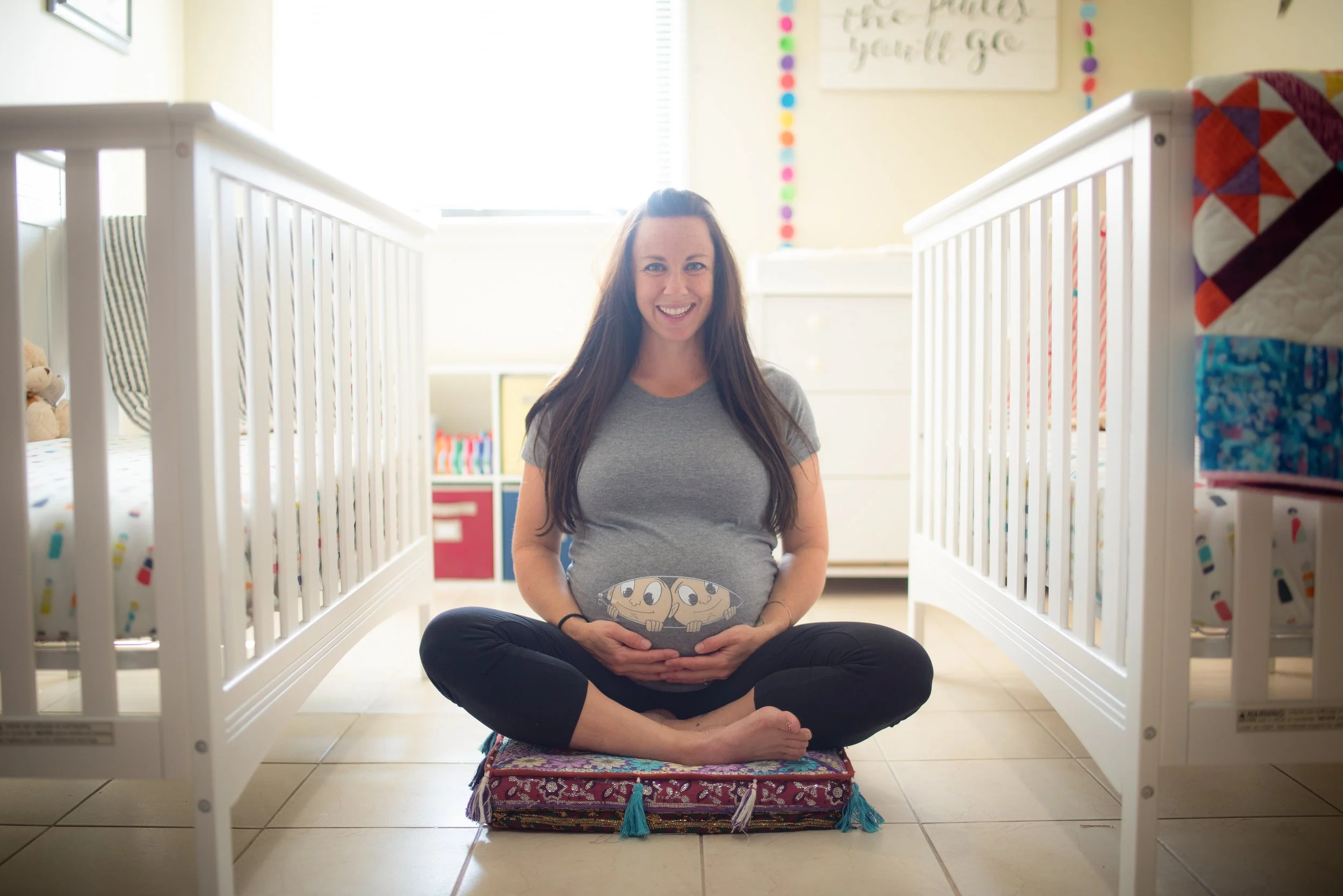 A pregnant woman sitting cross-legged on a colorful cushion between two white cribs in a brightly lit nursery, smiling and cradling her belly.
