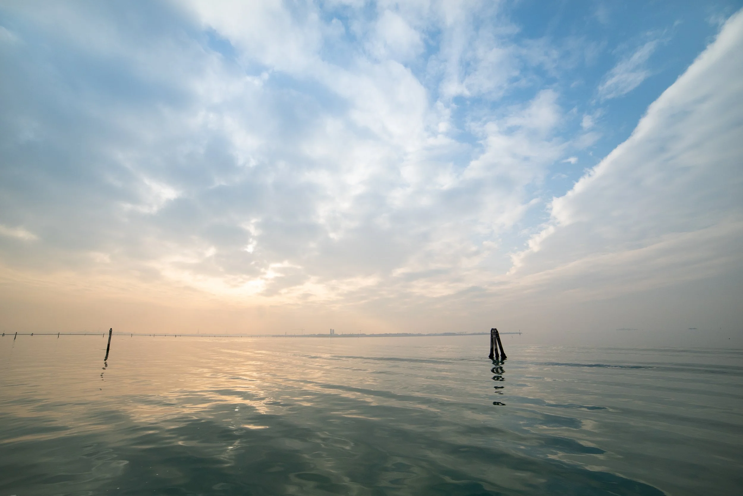 Calm water body with a few boat poles and a distant city skyline under a partly cloudy sky during sunrise or sunset.