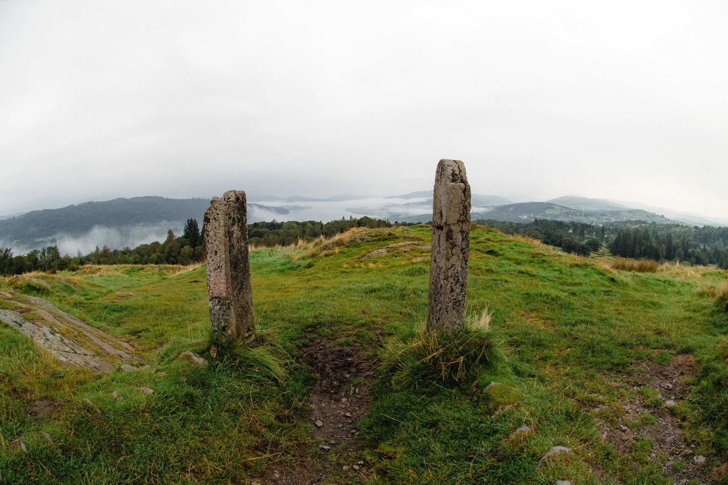 Two standing ancient stone markers on a grassy hill overlooking foggy rolling hills and a forested landscape.