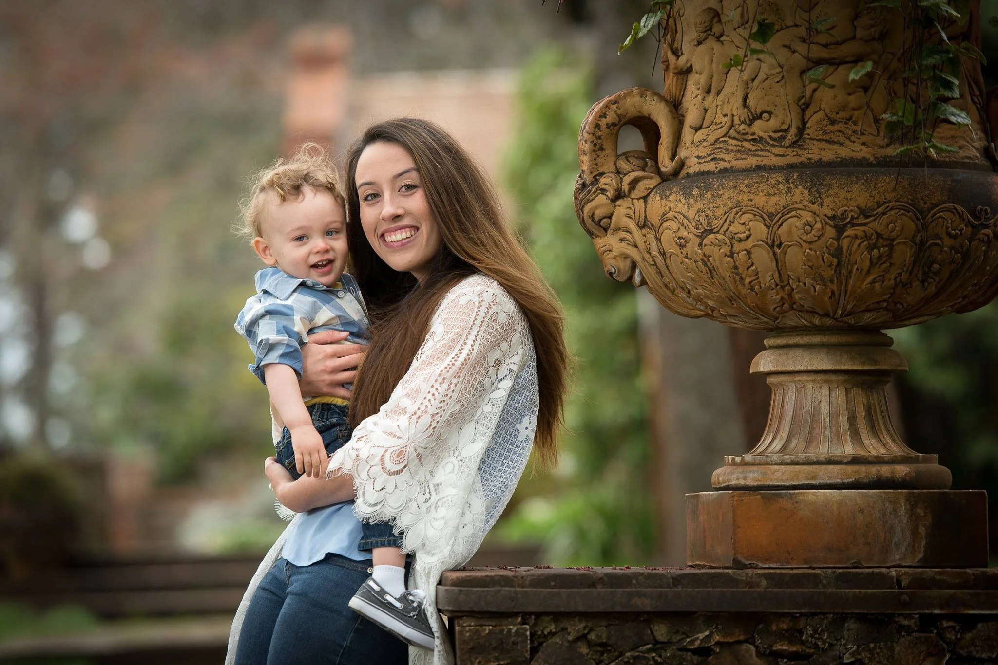 A smiling woman holding a young boy outdoors near a large ornate stone planter.