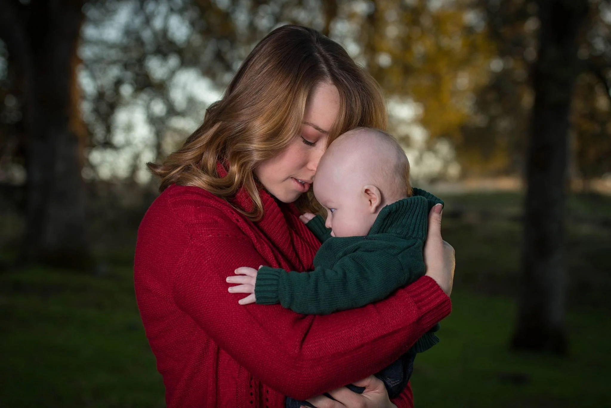 A woman with red hair wearing a red sweater holds a baby dressed in a green sweater in an outdoor setting with trees in the background.