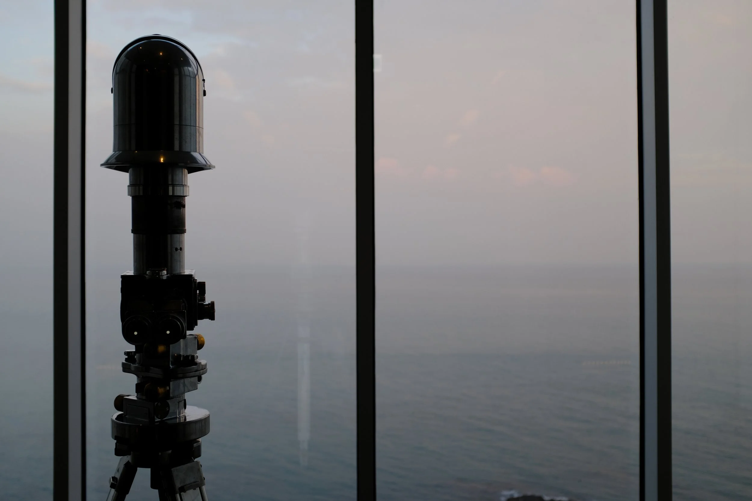 A telescope on a tripod located indoors, looking out through large window panels at a cloudy sky over the ocean.