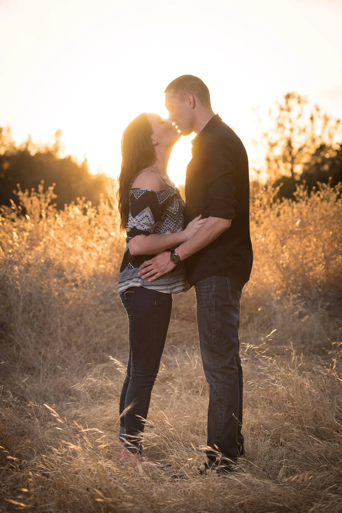 A couple standing close and about to kiss in a field at sunset, with tall dry grass and trees in the background.