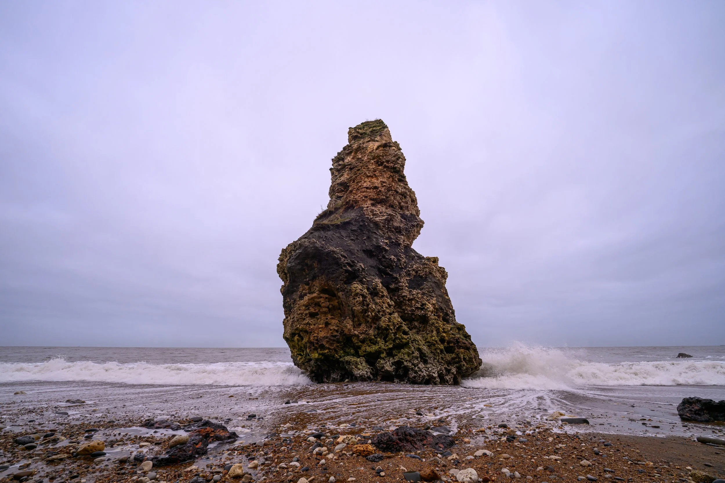 A large, irregularly shaped rock formation rising from the sandy beach near the ocean, with waves crashing at its base and an overcast sky overhead.