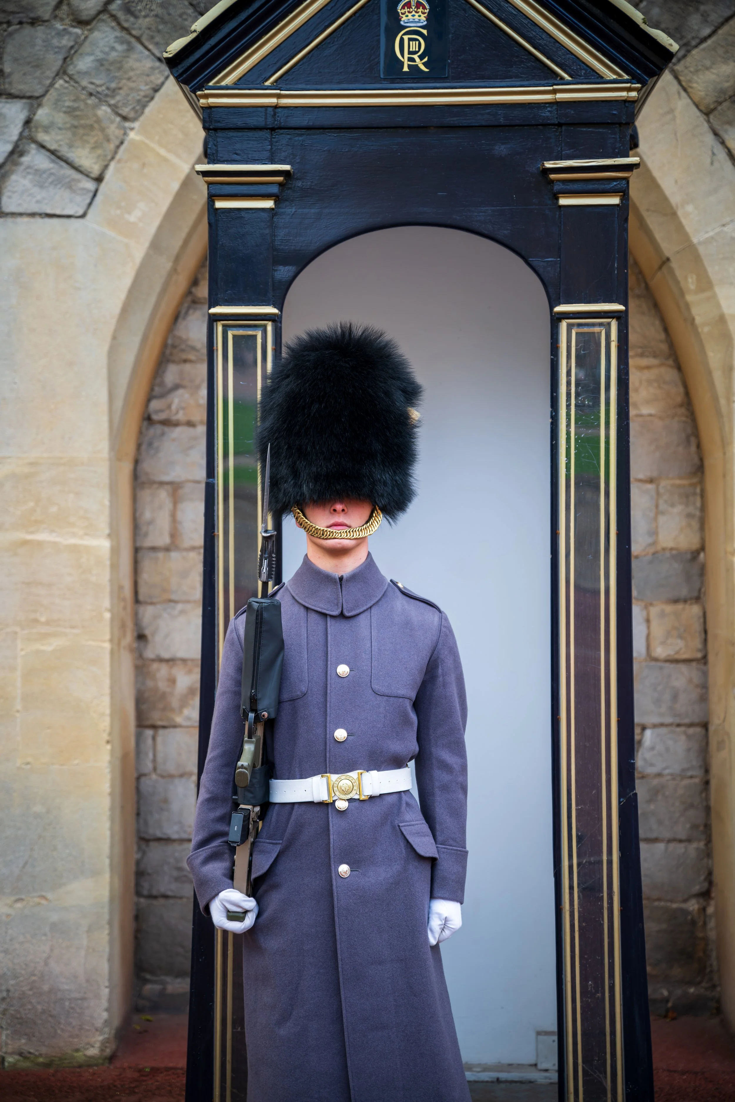 A British royal guard in a gray uniform with a tall black bearskin hat stands in front of a stone wall, holding a rifle.