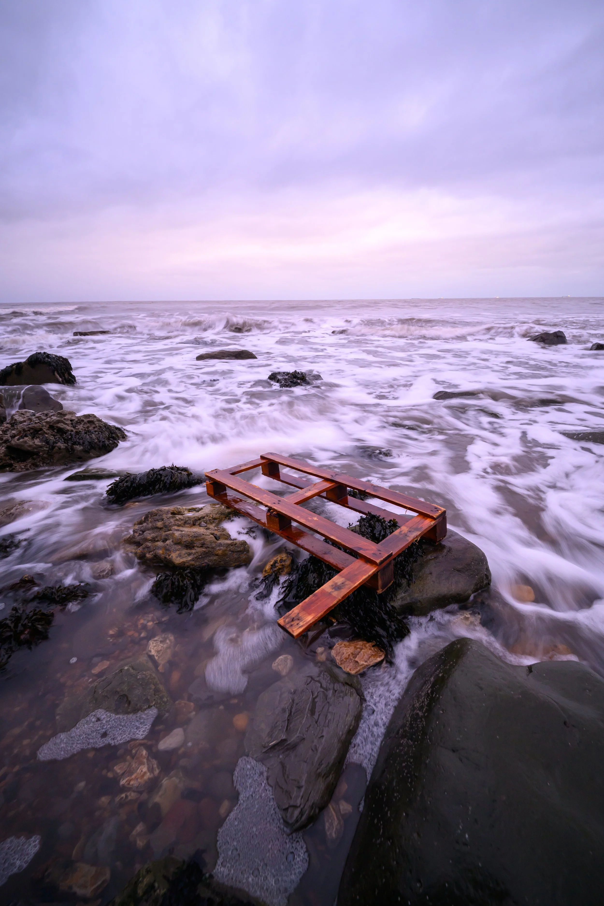 A wooden pallet lying on rocks at the edge of the ocean with waves crashing against it, under a cloudy sky.