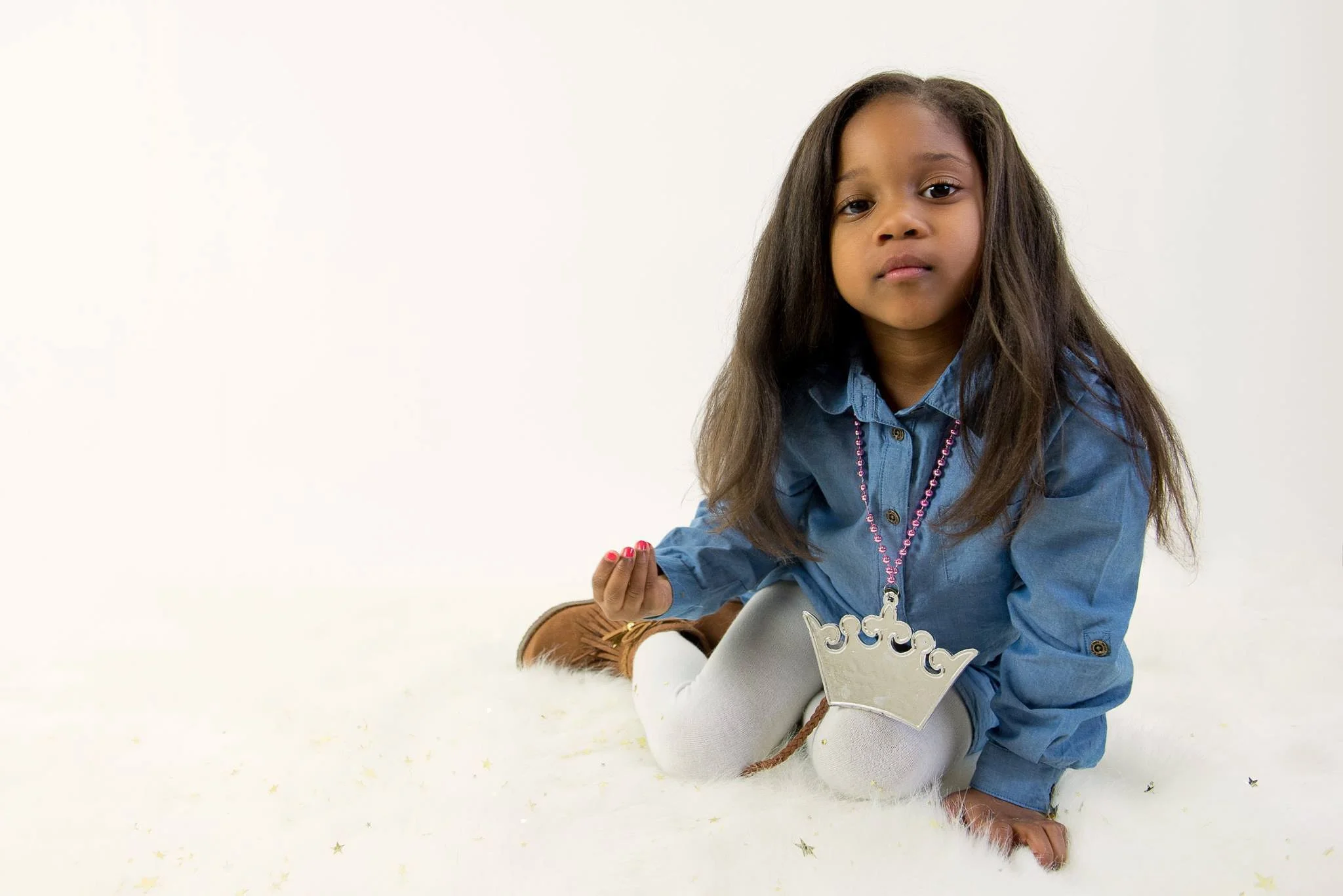 Young girl with long, dark hair, wearing a blue denim shirt, white tights, and brown boots, sitting on a white fluffy surface, holding a silver crown necklace.