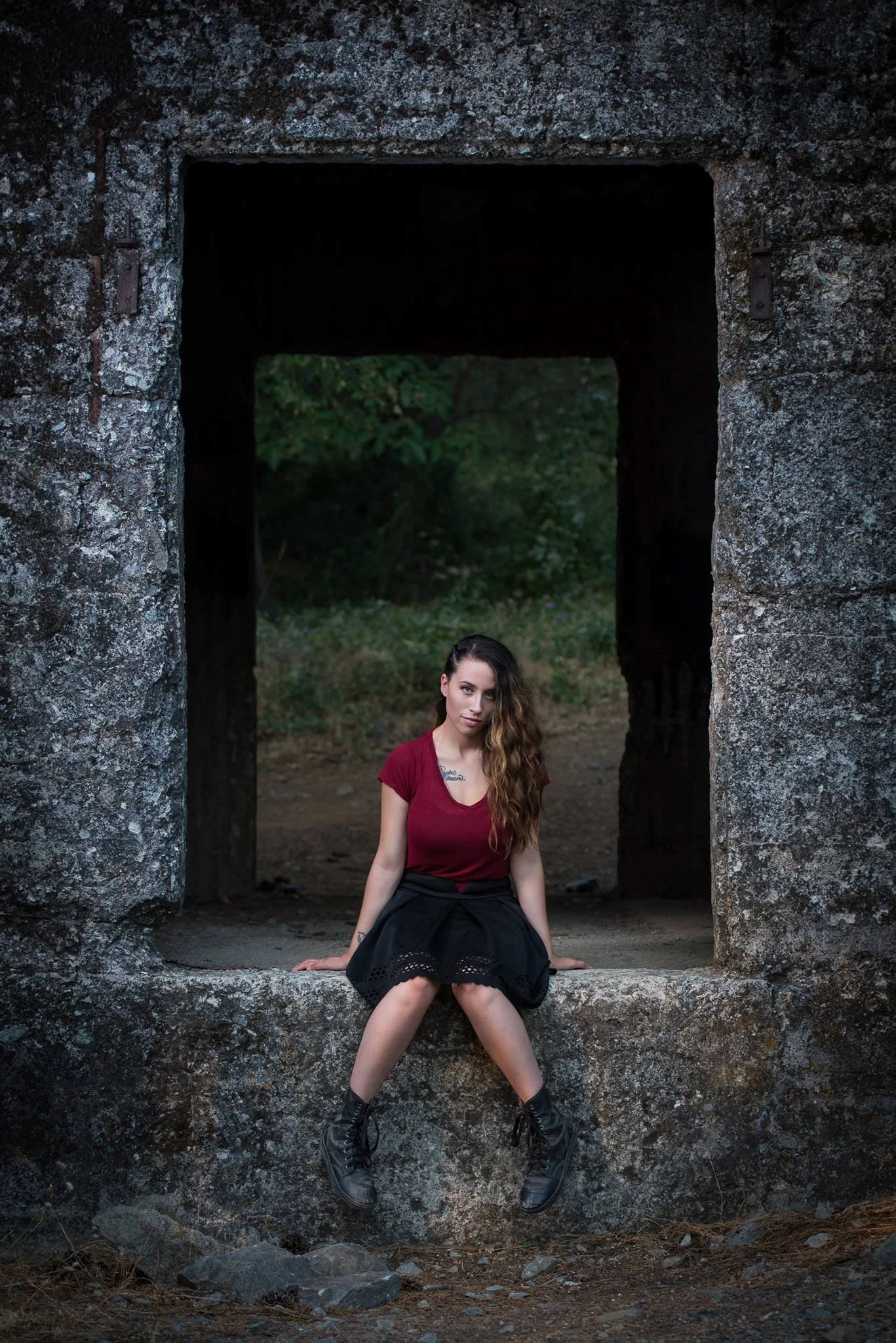 A young woman with long wavy hair, wearing a red t-shirt, black skirt, and combat boots, sits on a stone ledge in front of a large stone archway with rustic, textured walls, surrounded by greenery.