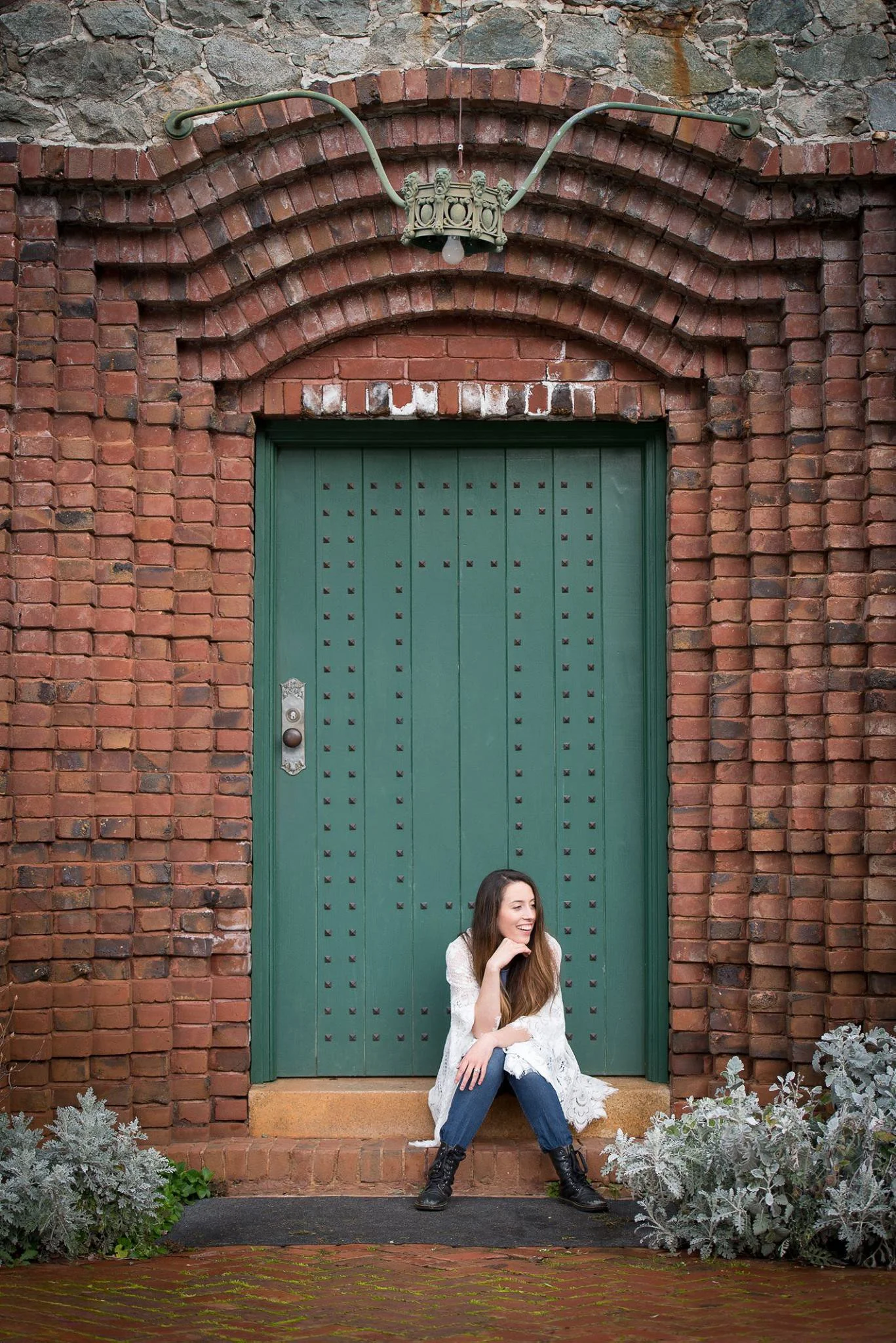 A young woman with long brown hair, wearing a white lace cardigan, blue jeans, and black boots, sits on a brick step in front of a large green wooden door framed by red brick archways. She is smiling and looking to her left, with greenery and plants 