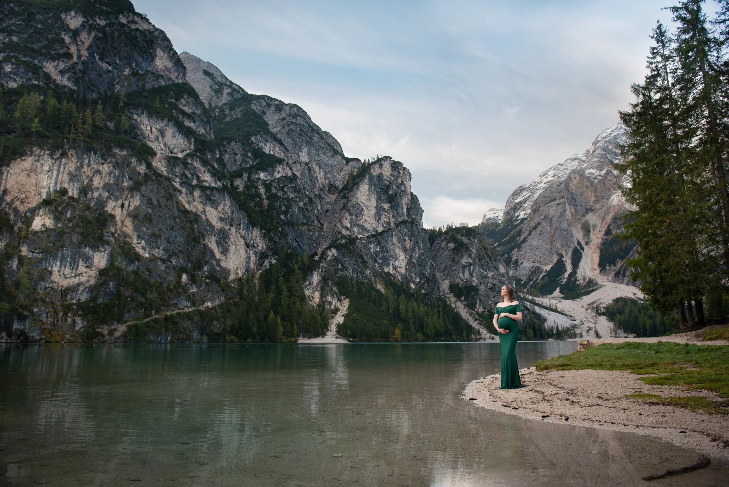 A pregnant woman in a green dress stands on the edge of a lake in a mountainous landscape with snow-capped peaks, green trees, and cloudy sky.