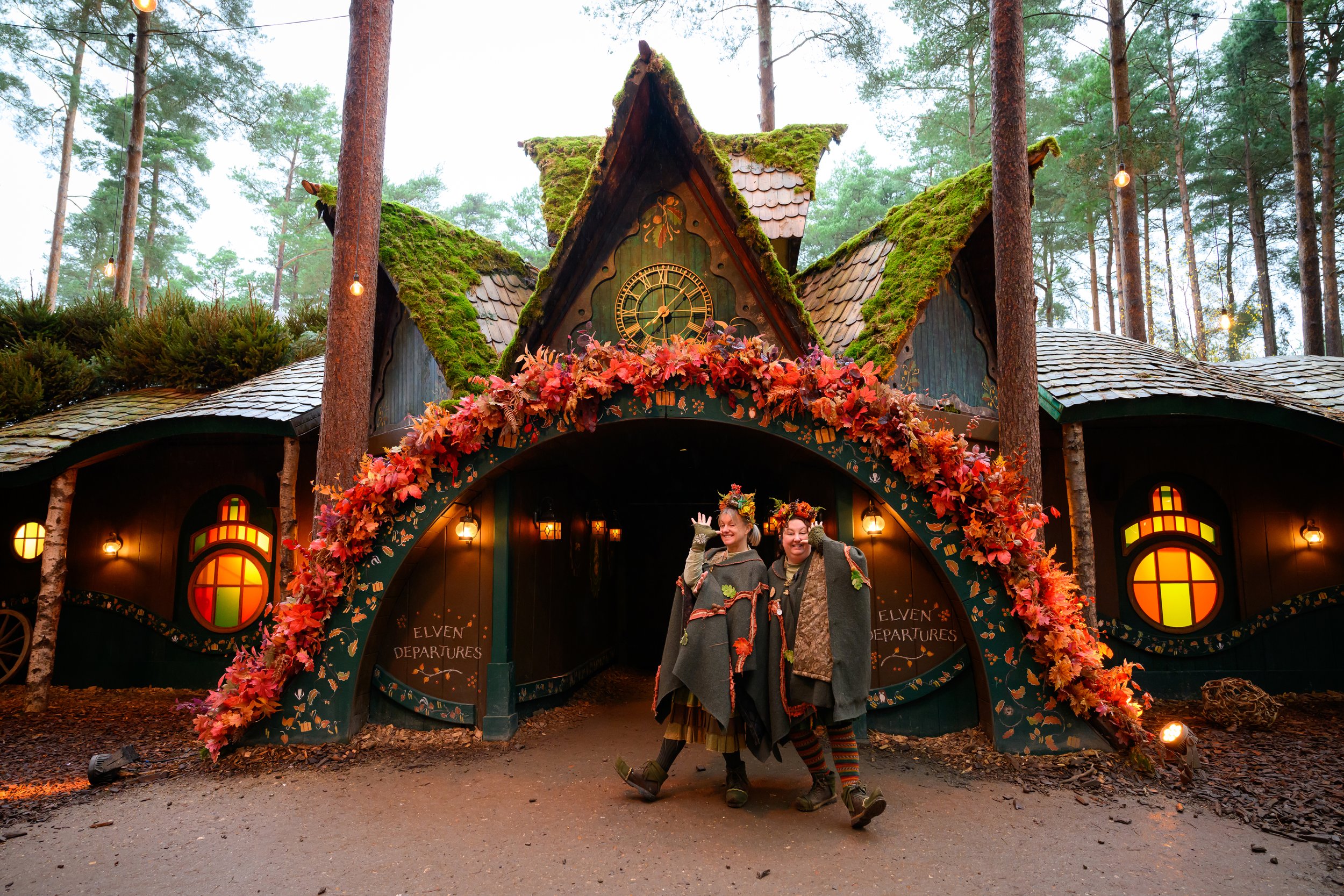Two people dressed as elves in costumes standing and smiling in front of a whimsical, fairy-tale like building decorated with autumn leaves and moss, surrounded by trees.