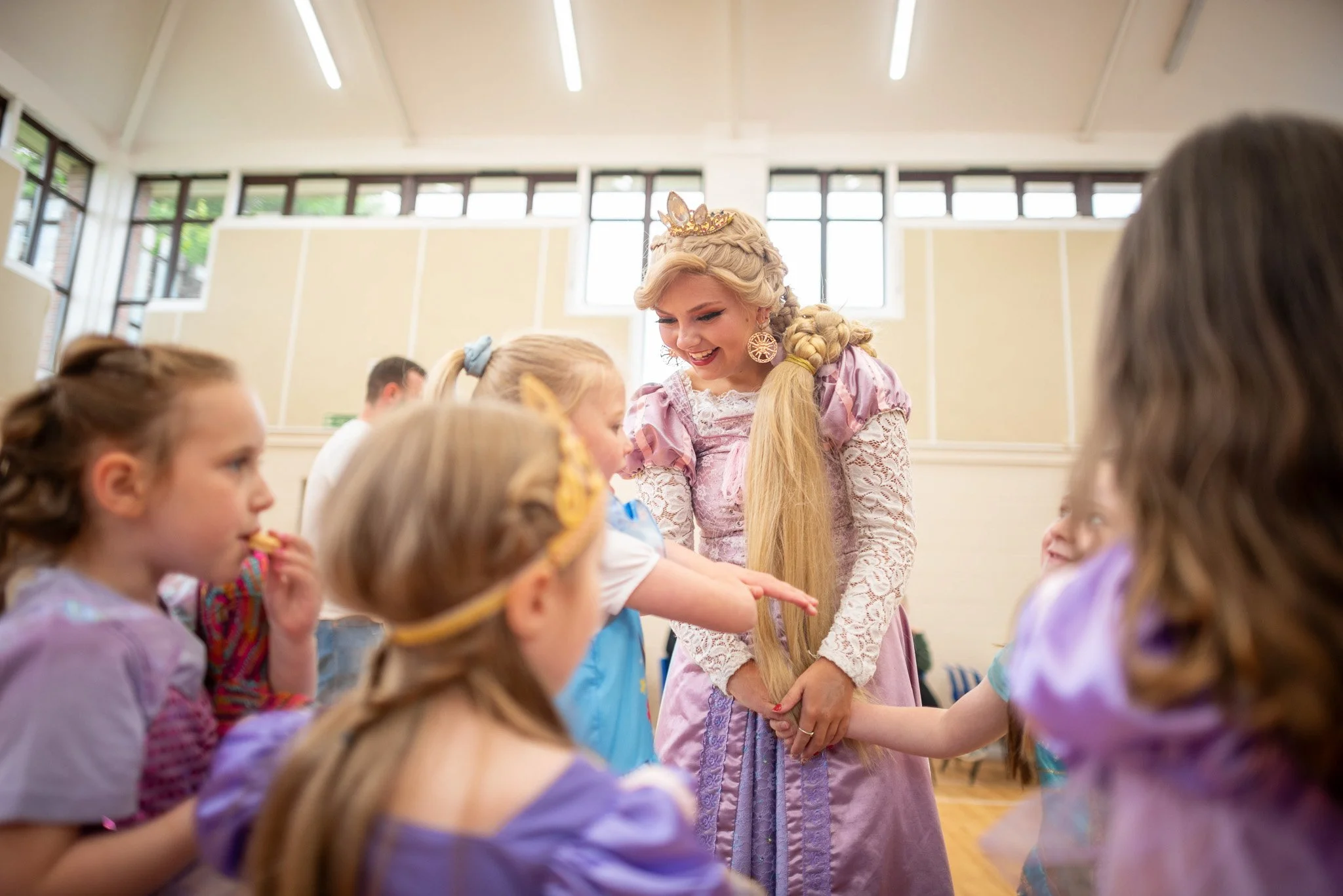 A woman dressed as a princess with long blonde hair, a tiara, and a pink gown, is smiling and interacting with a group of young girls dressed as princesses at a children's party.
