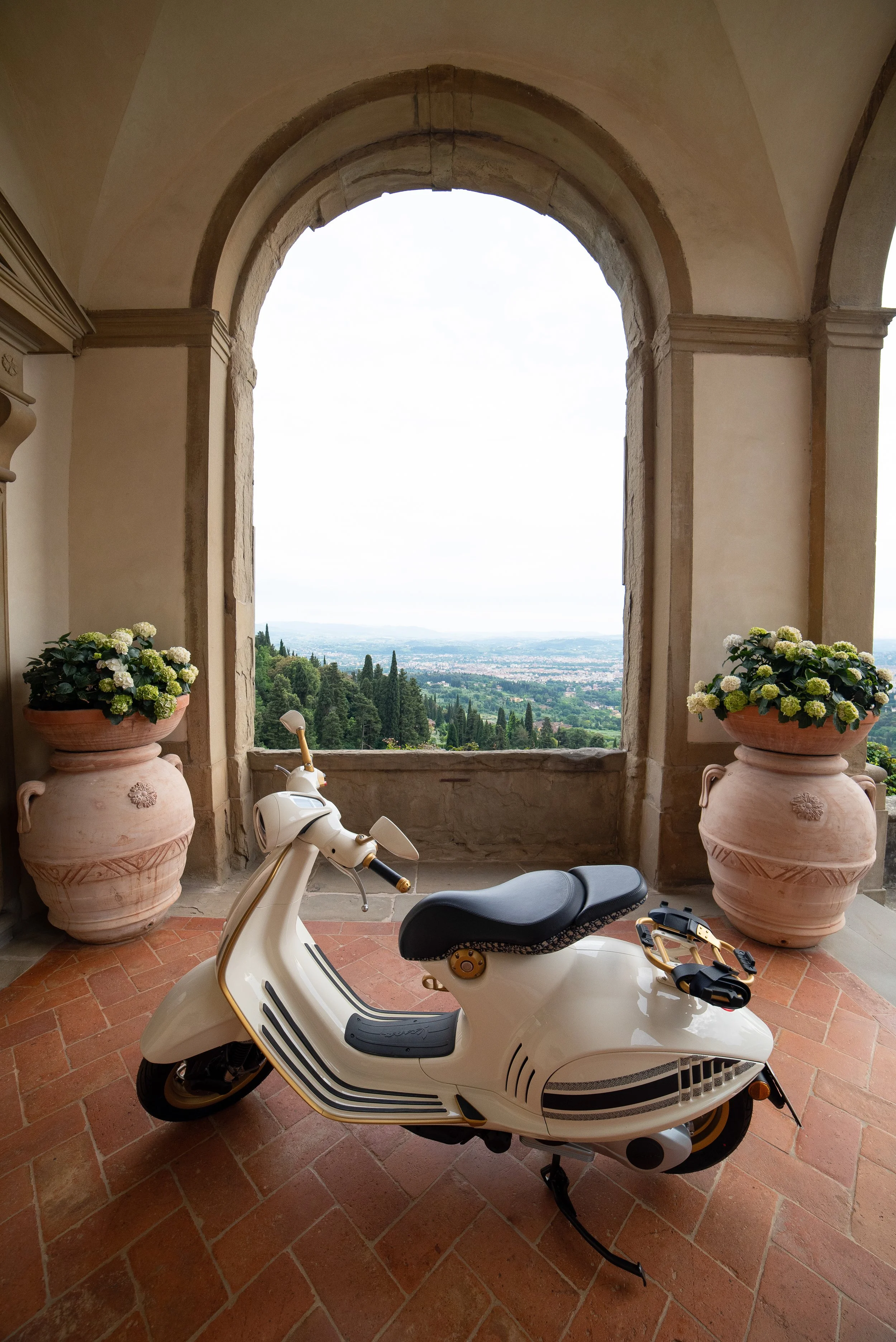 A vintage white scooter parked on a terracotta tiled floor inside a castle or historic building with large open archways and potted flowers, overlooking a lush green landscape with trees and distant city view.