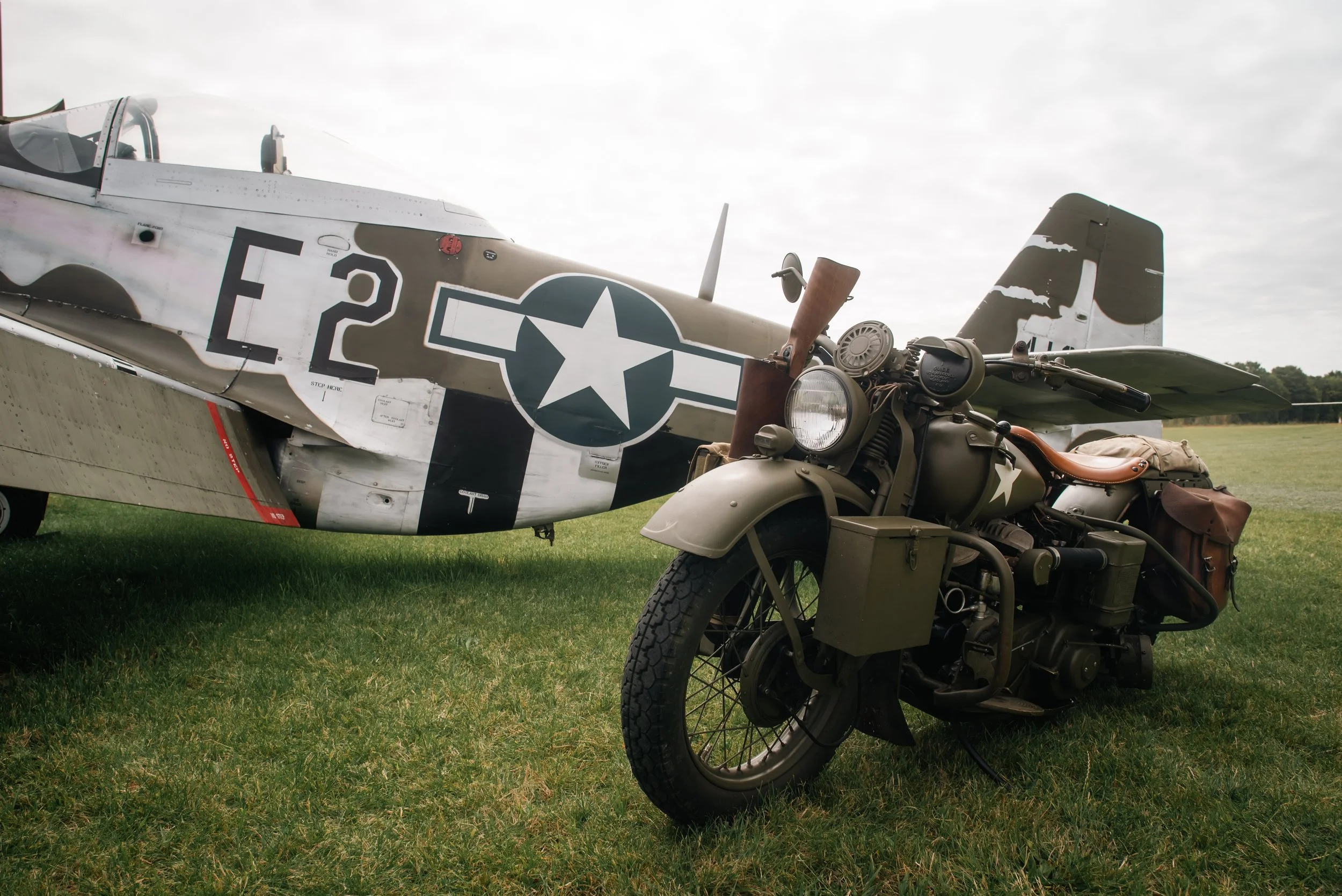 A military aircraft and a vintage motorcycle parked on a grassy field under an overcast sky.