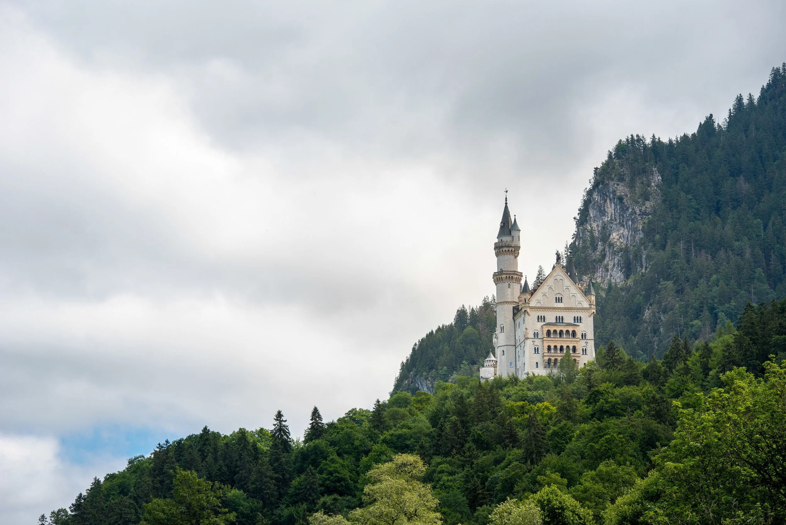 A castle on a hill surrounded by dense green trees, with mountains in the background and a cloudy sky above.