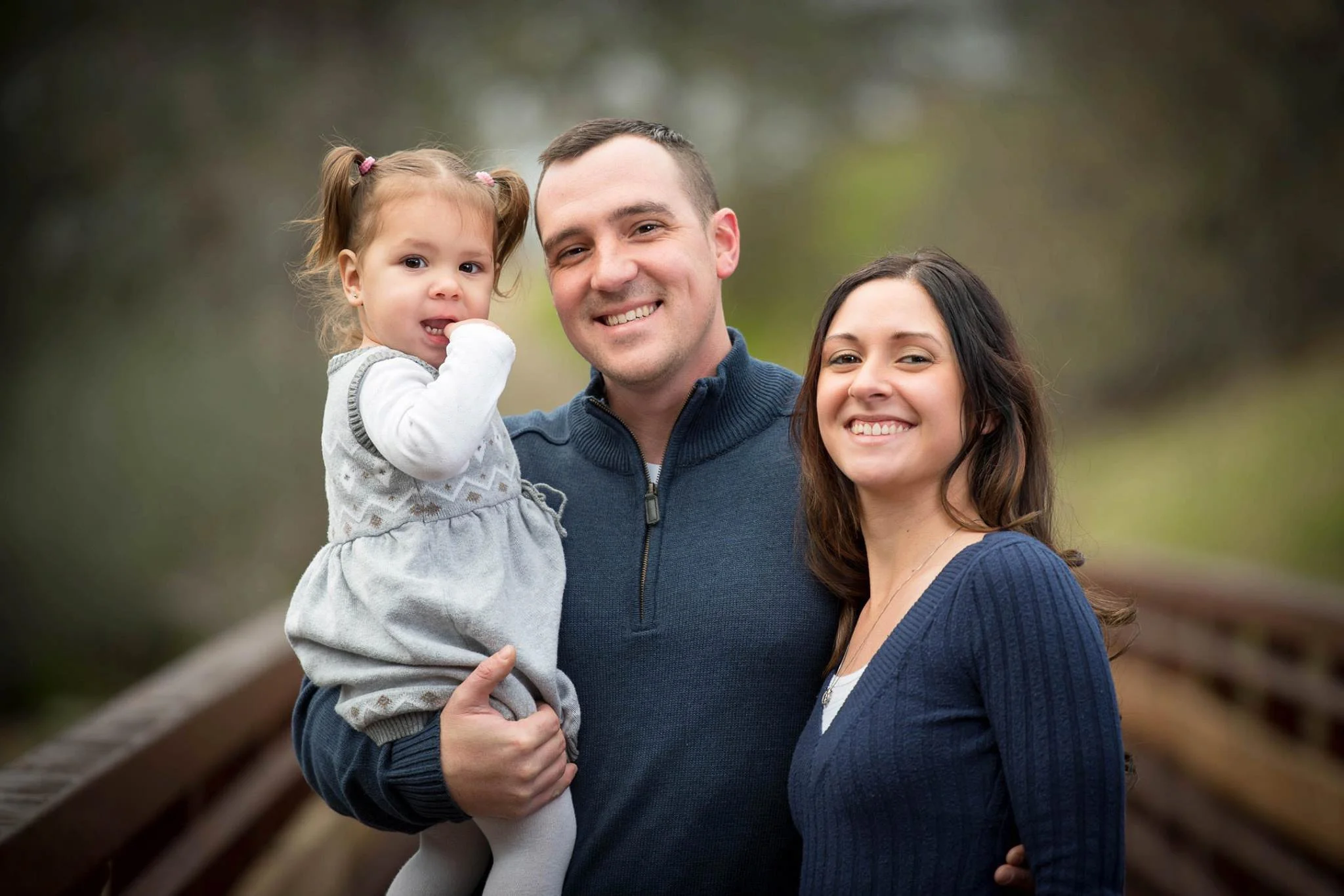 A happy family of three, a man, a woman, and a young girl, standing together outdoors on a wooden bridge with trees in the background. The man is holding the girl, and they are all smiling at the camera.