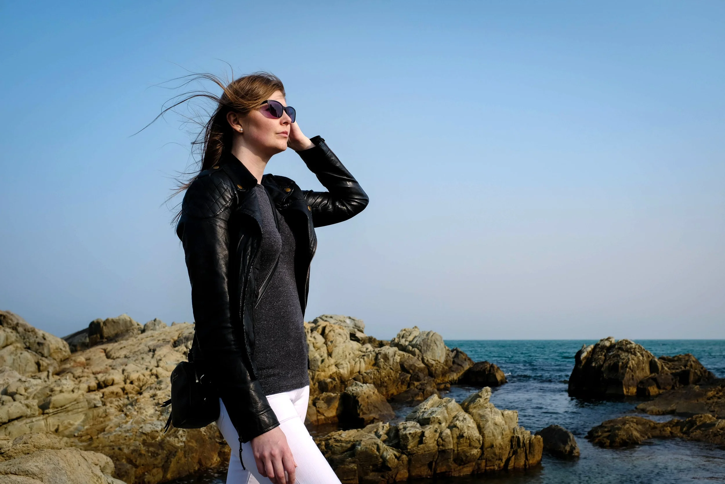 A woman wearing sunglasses, a black leather jacket, a gray shirt, and white pants stands on rocky shore by the ocean, facing left with her hand near her hair.