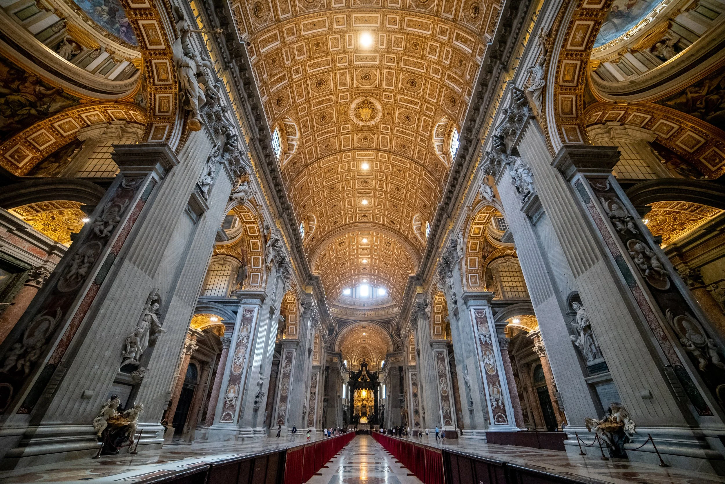 Interior of a grand, ornate cathedral with tall marble columns, intricate gold detailing on the ceiling, and a central aisle leading to an altar with a large gilded sculpture.
