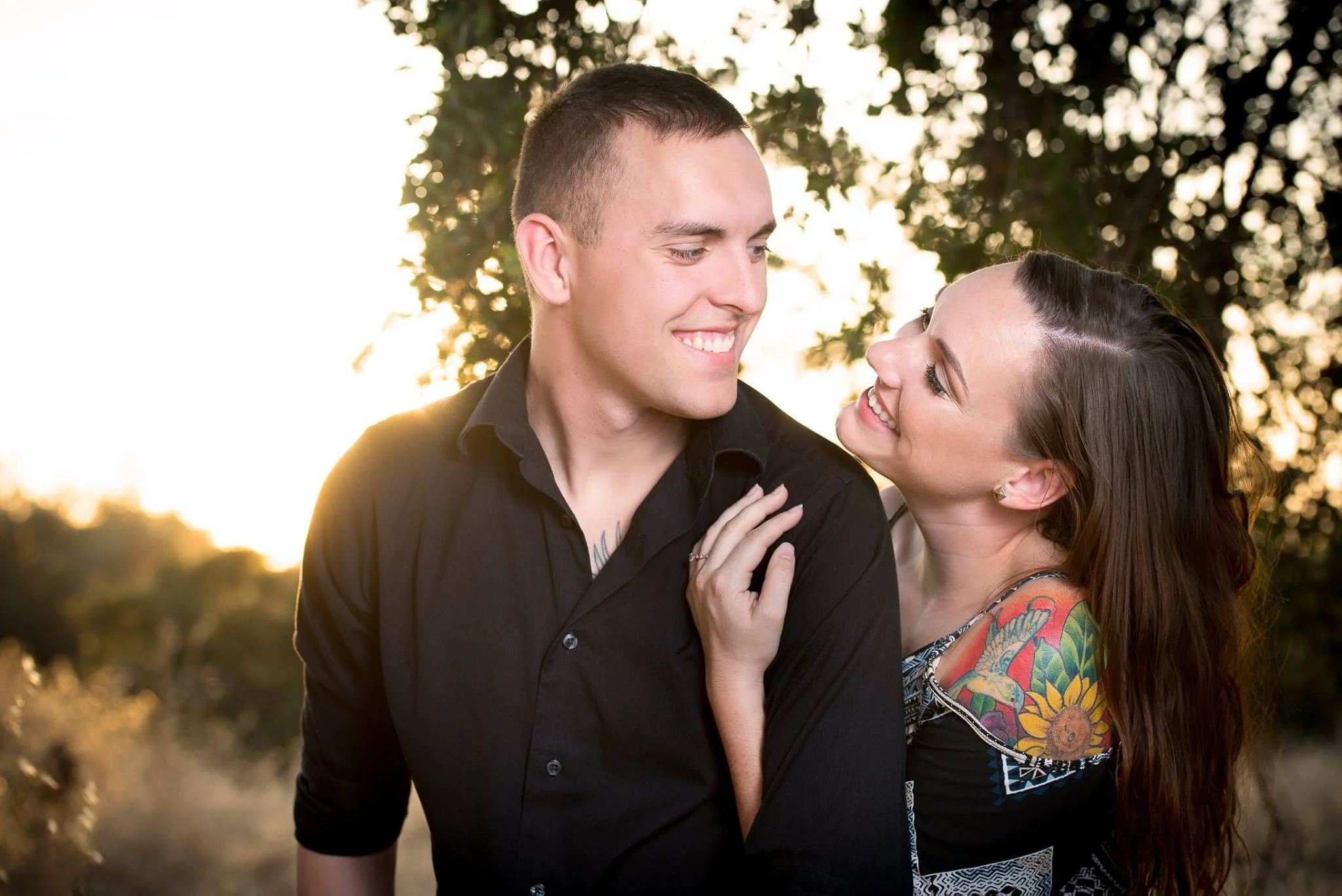 A smiling couple outside during sunset, with the woman looking at the man and showing colorful tattoos on her shoulder.