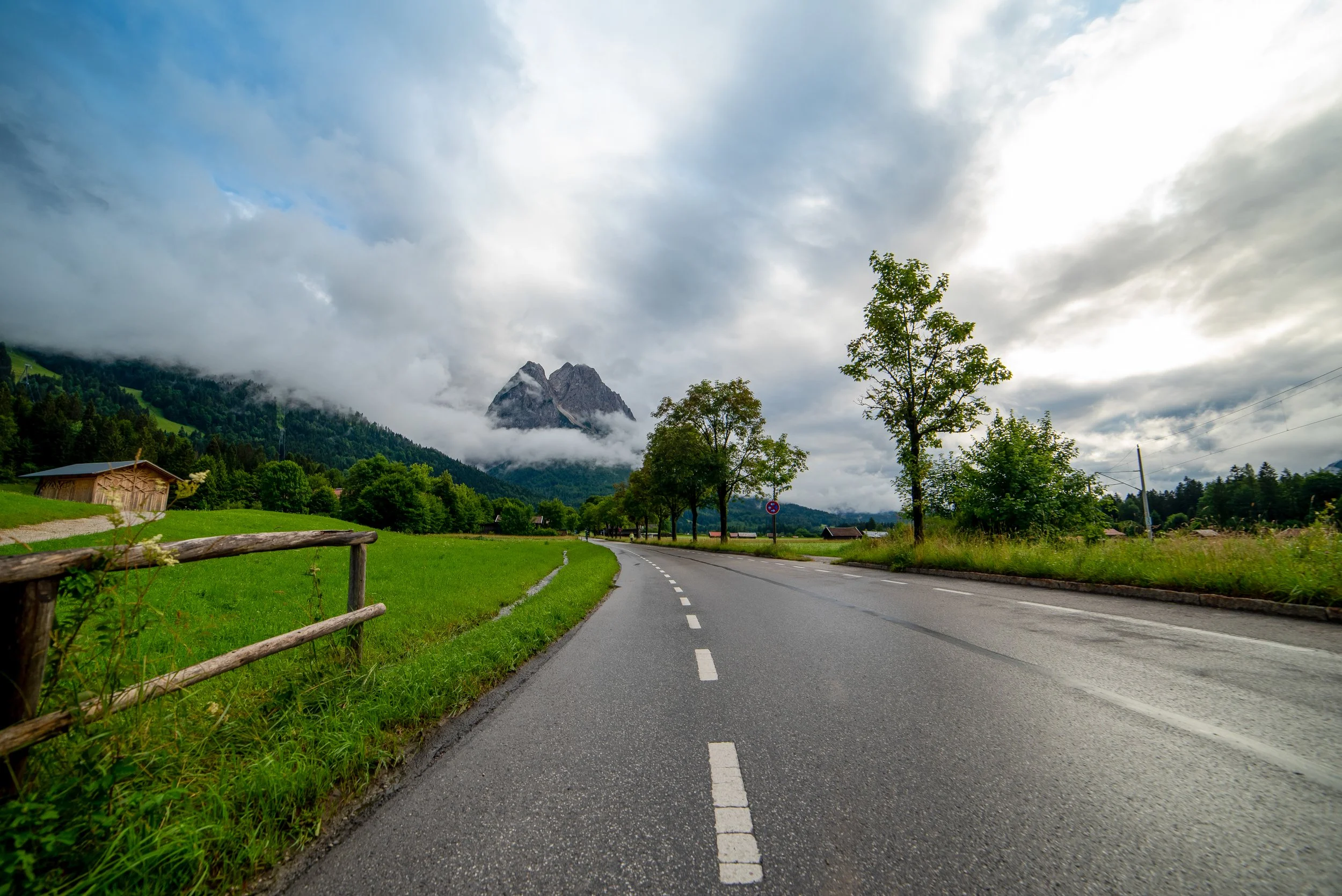 A winding road through a lush green landscape with trees and distant mountains partly covered in clouds.